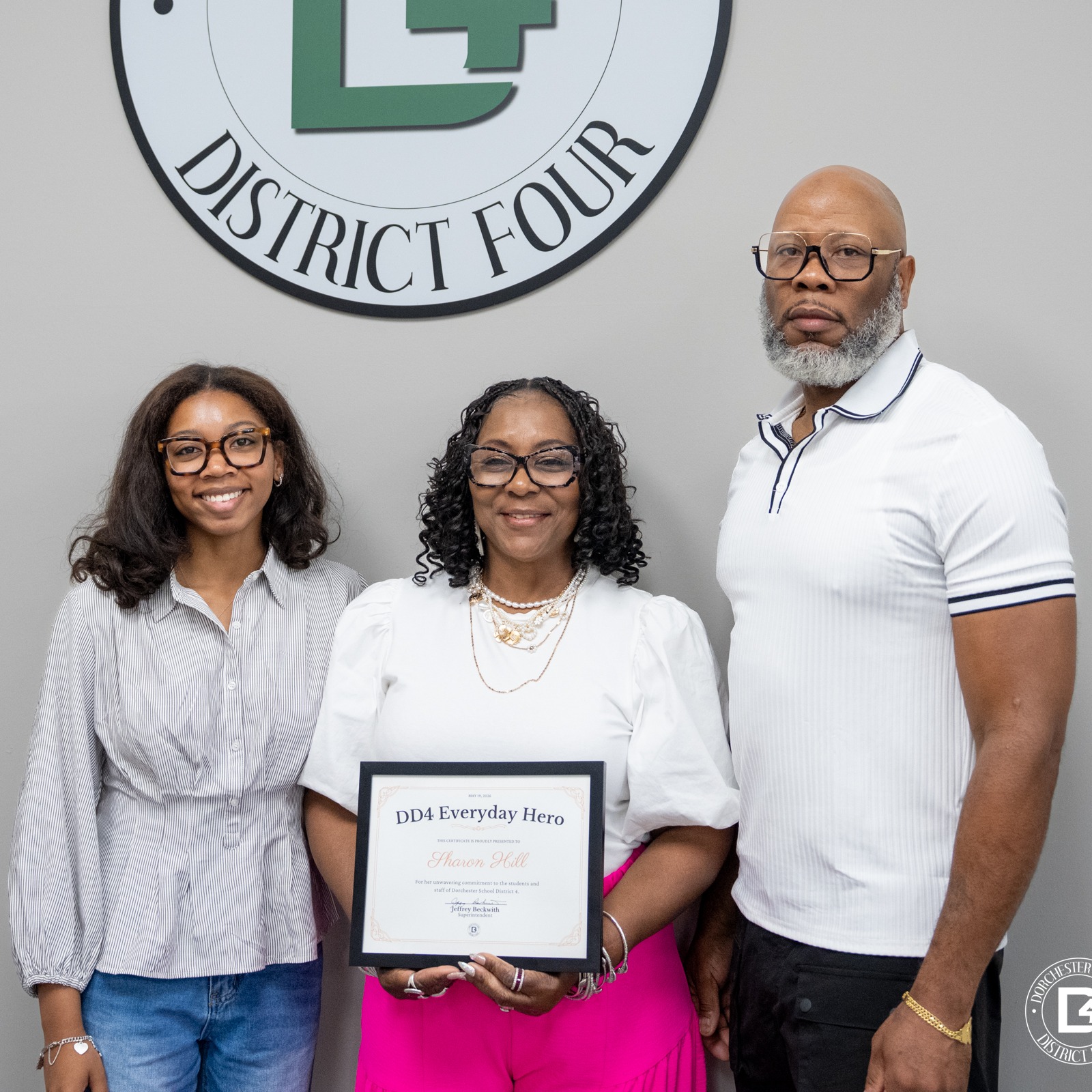 Sharon Hill stands with two family members beneath the Dorchester School District Four logo while holding a DD4 Everyday Hero certificate. A young woman stands beside her smiling in glasses and a striped blouse, while a man in a white polo stands on the other side posing for the photo.