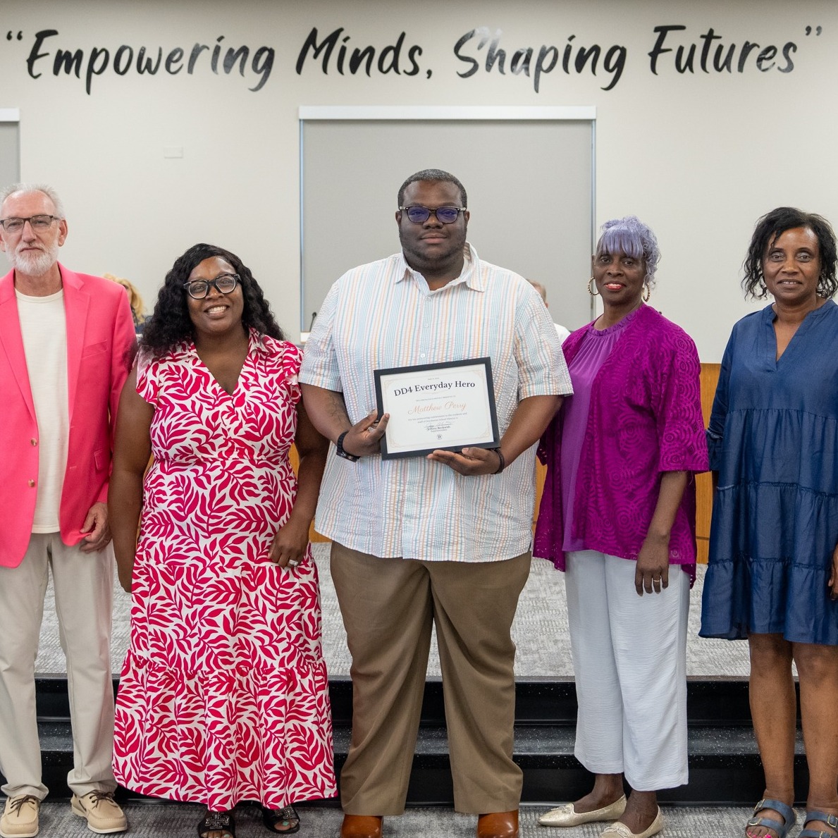 Matthew Perry stands in the Dorchester School District Four board room holding a DD4 Everyday Hero certificate while posing with Ms. Hughes, Mr. Earle, Mrs. Britt, Mr. Beckwith, and co-workers. The group stands beneath the district’s “Empowering Minds, Shaping Futures” slogan for a recognition photo.