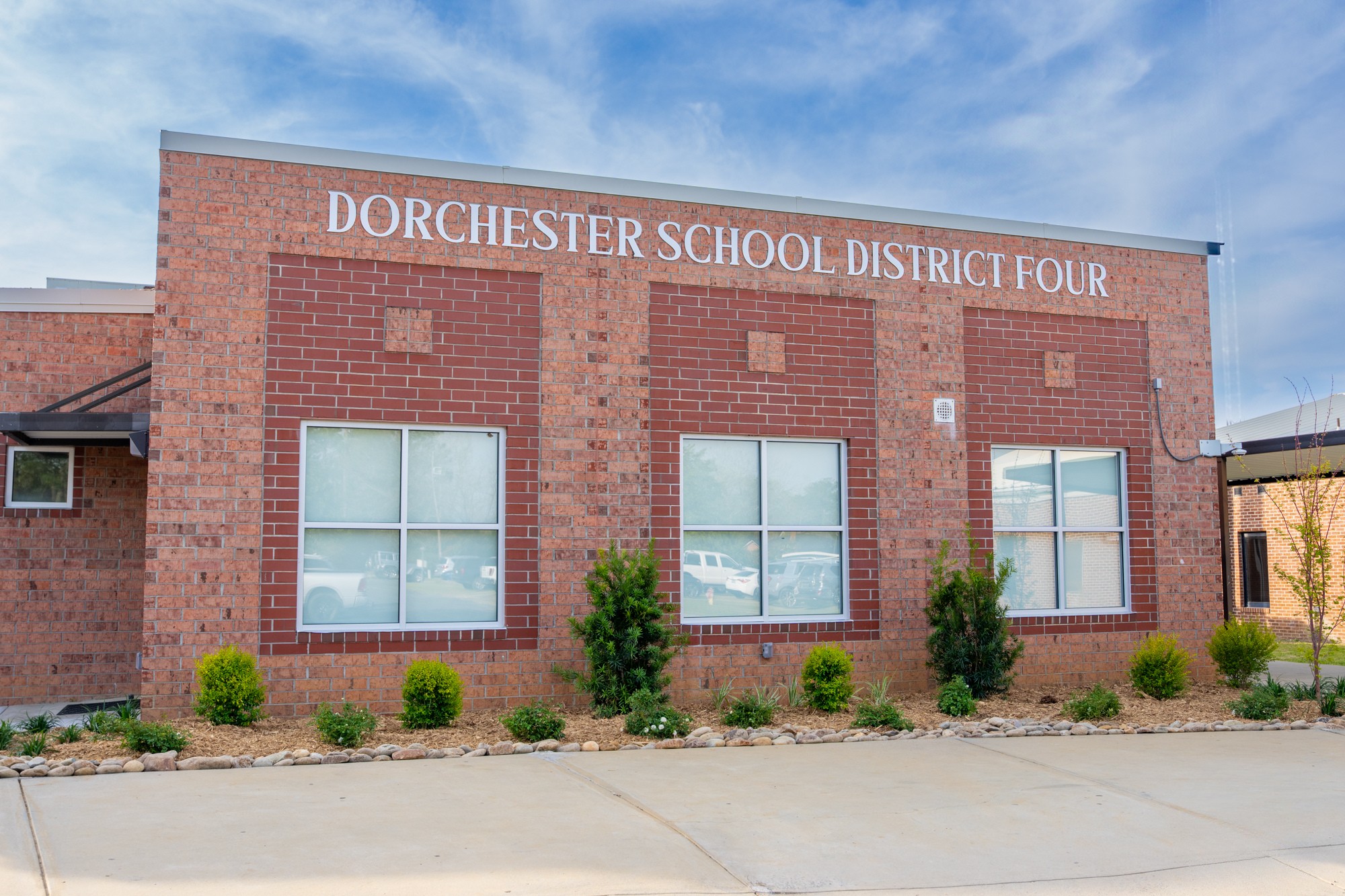 Exterior of the Dorchester School District Four administrative building with the district name displayed across the brick facade, landscaped shrubs and trees in front, and a blue sky with wispy clouds overhead.