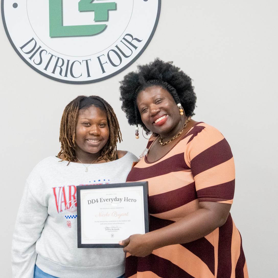 Nicole Bryant posing with her daughter in front of the district logo, holding her DD4 Everyday Hero Certificate