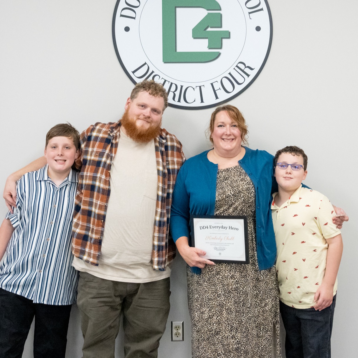 Mrs. Chubb posing with her husband and two male children in front of the DD4 logo on a wall. Mrs. Chubb is holding her Everyday Hero Certificate.