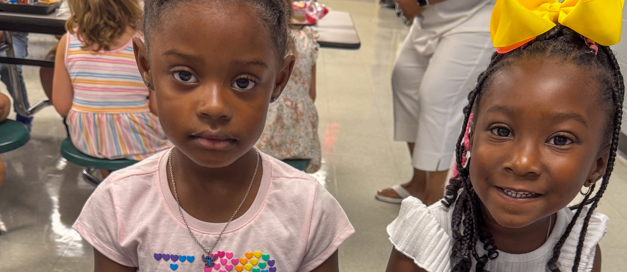 Two young students from Clay Hill Elementary School sit side by side at a cafeteria table during breakfast. One girl smiles brightly while the other looks calmly at the camera. Both are wearing colorful bows and t-shirts with cheerful designs. A breakfast tray and milk carton are visible on the table. The Dorchester School District Four logo is shown in the lower right corner.