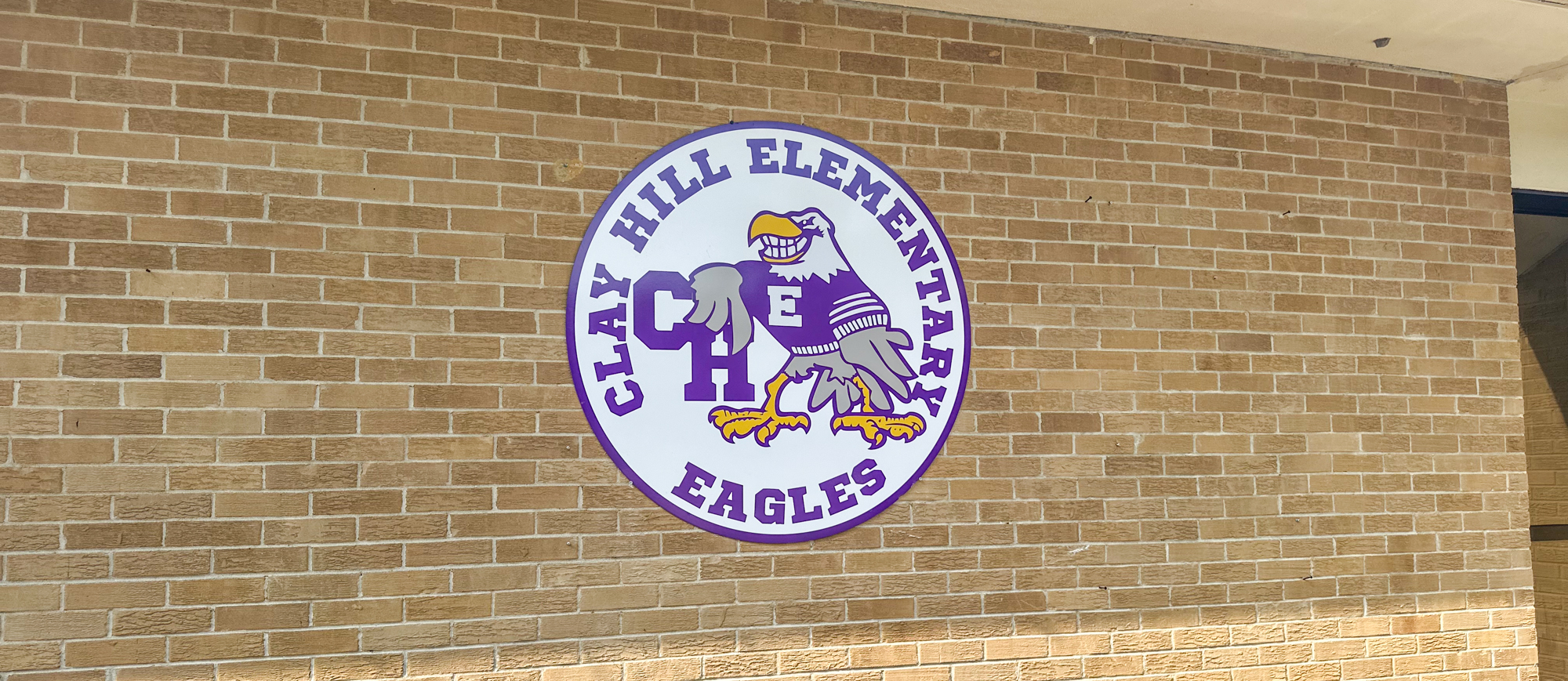 The Clay Hill Elementary School logo displayed on a brick wall. The circular logo features a purple and gold eagle wearing a sweater with the letter "C," and the words "Clay Hill Elementary Eagles" surrounding it.