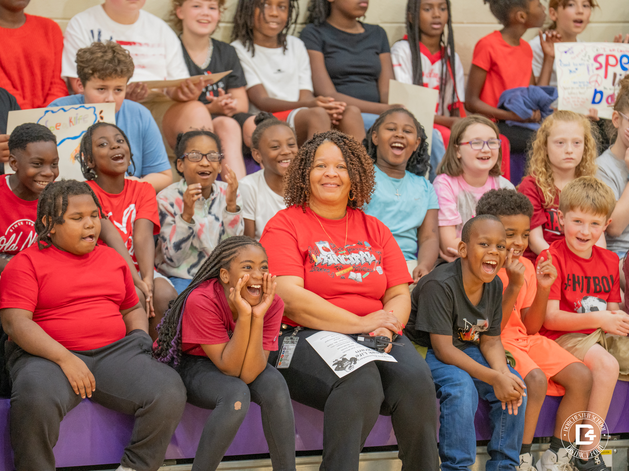 Clay Hill Elementary Principal Alisa Lemon sits in the bleachers surrounded by smiling and laughing students during the 2nd annual Speak Life Day event. Students wearing red, black, and white shirts cheer and shout with excitement, holding posters in the background. The joyful expressions capture the uplifting spirit of the day.