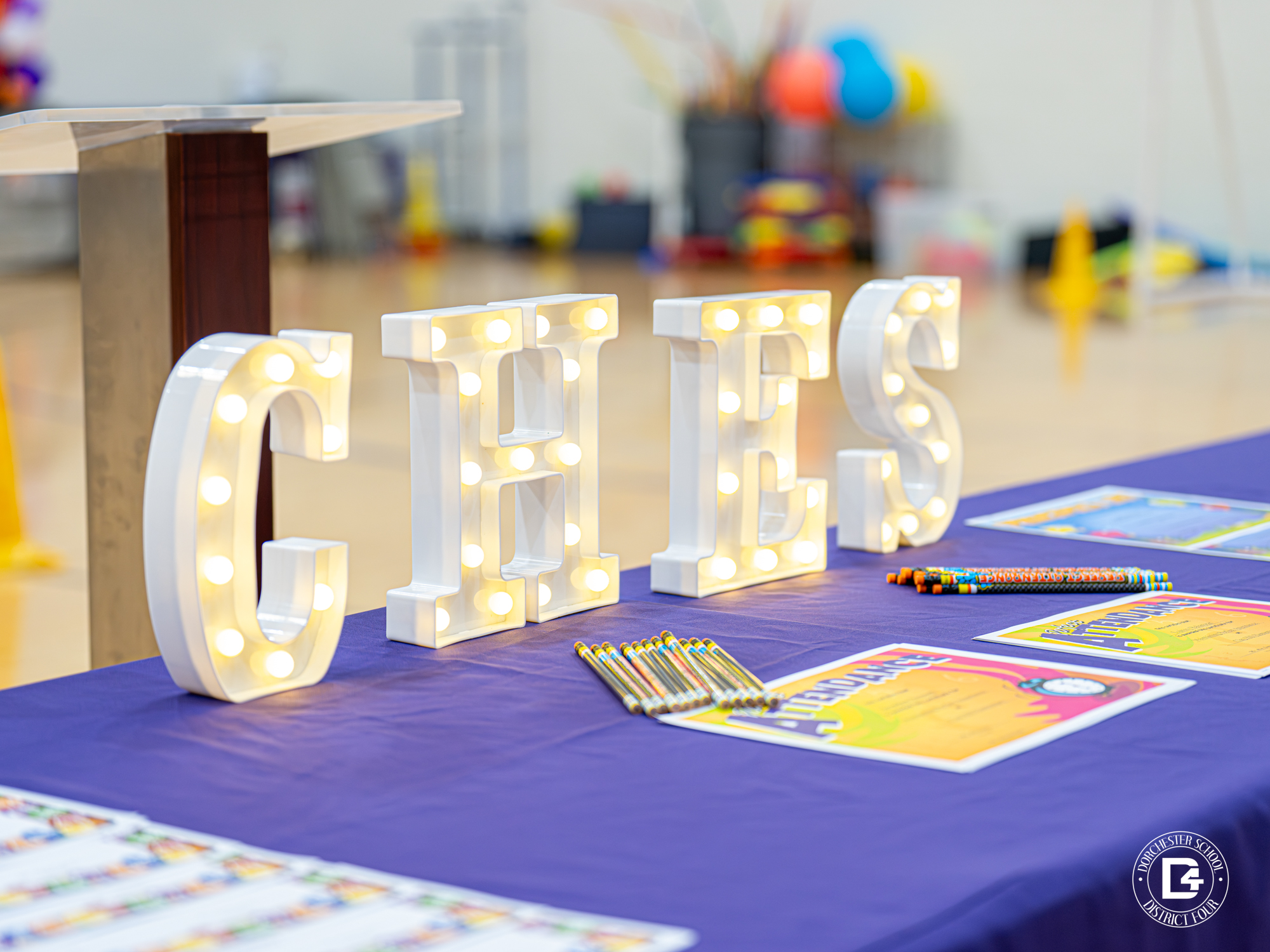 A close-up of a decorated table covered in a purple cloth with large white marquee-style letters spelling “CHES.” The table holds colorful pencils, certificates, and papers for an event inside a gymnasium. The background shows blurred gym equipment and decorations.