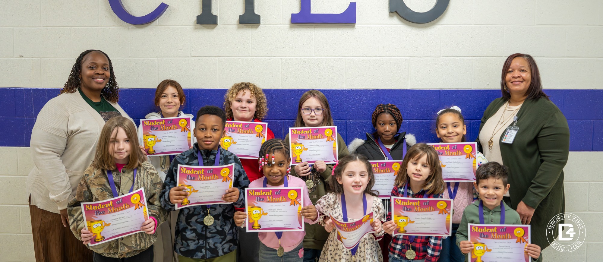Group photo of Clay Hill Elementary Students of the Month standing in front of a wall with large “CHES” letters, holding certificates and medals, with two adult staff members standing at each end of the group.