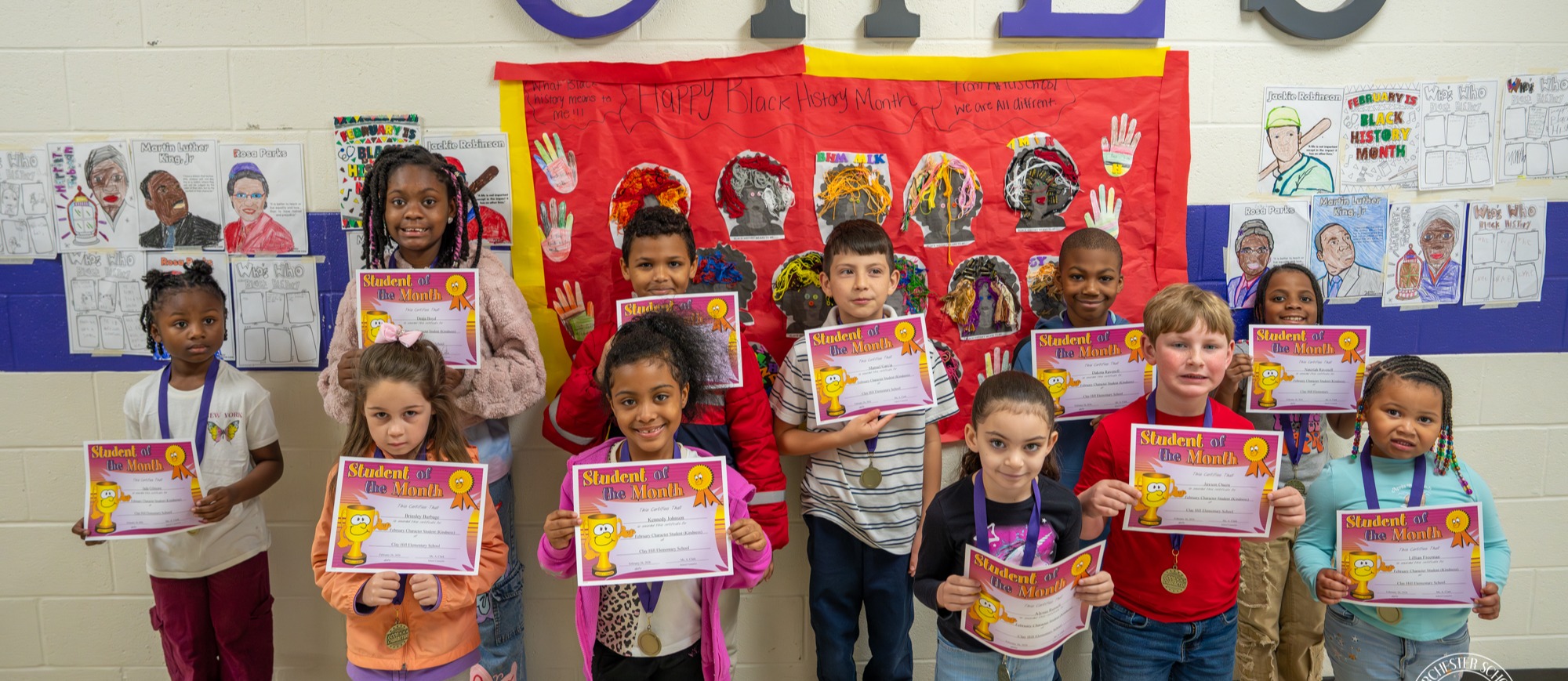 A group of elementary students stand in a school hallway under large “CHES” letters, holding “Student of the Month” certificates and wearing medals. Behind them is a red “Happy Black History Month” display board with student artwork and portraits. The students were recognized at Clay Hill Elementary School for demonstrating kindness.