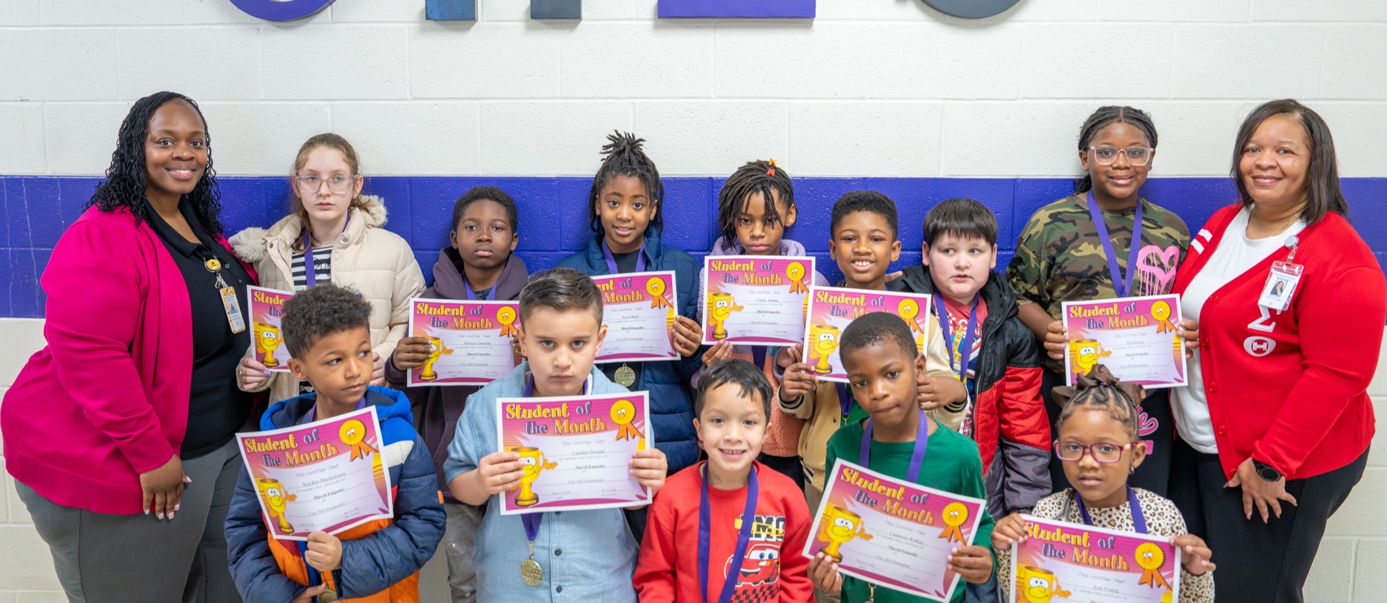 A group of Clay Hill Elementary School students stand in front of a wall with large “CHES” letters, each holding a “Student of the Month” certificate. The students, wearing medals, are joined by two staff members on either side, smiling and celebrating their achievement. The group represents a diverse set of young learners recognized for their hard work and positive behavior.