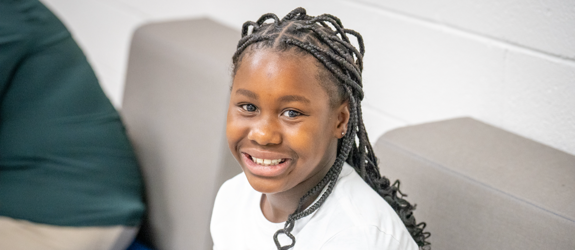 Smiling student with braided hair sits on a gray cushioned bench at Harleyville Elementary School, wearing a white T-shirt with a colorful graphic design.
