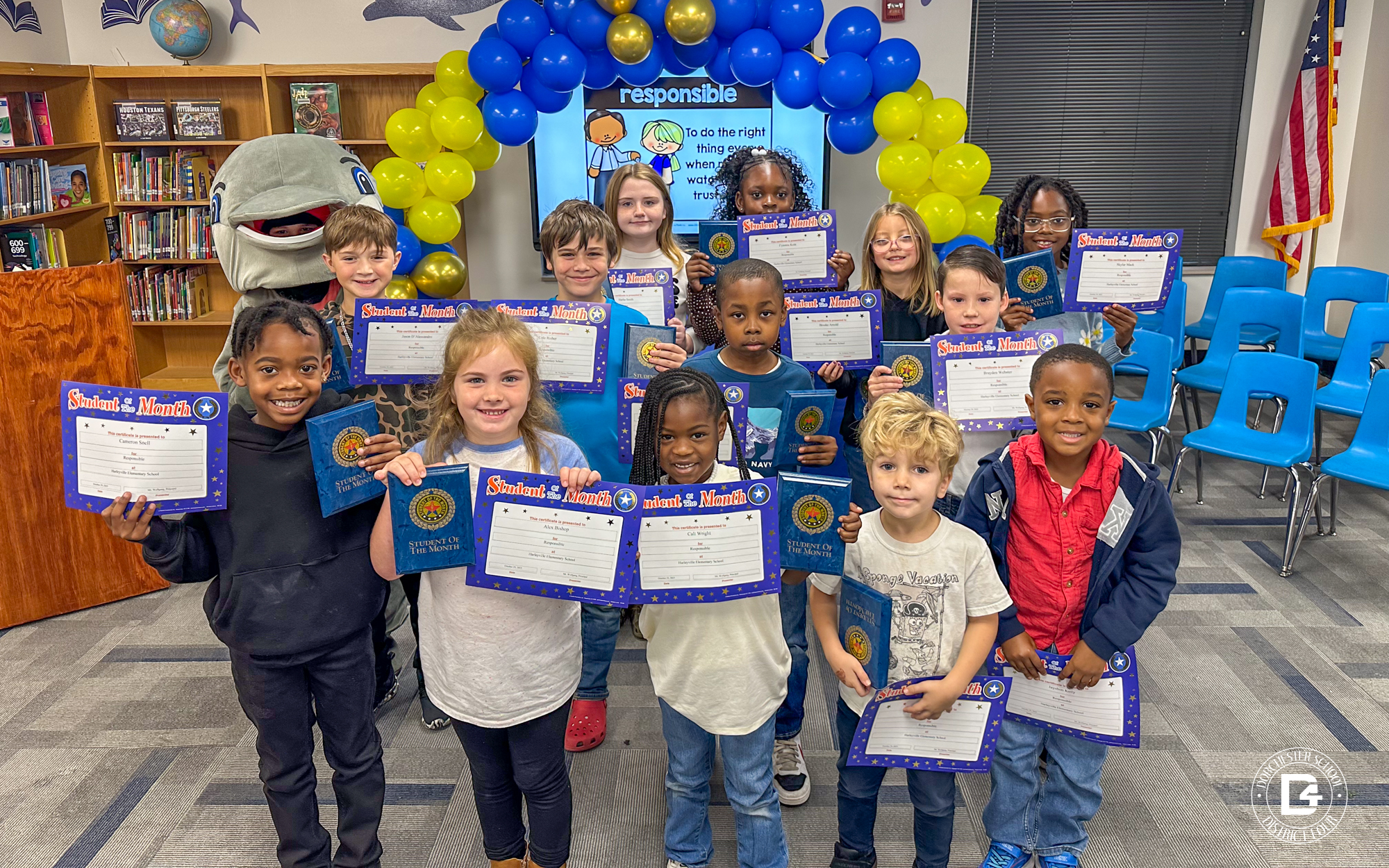 A group of smiling elementary students stand together in a library holding their “Student of the Month” certificates and blue award booklets. Behind them, a person in a dolphin mascot costume poses beside a balloon arch made of blue and yellow balloons. A screen in the background displays the word “responsible” with a short definition. The setting includes blue chairs, bookshelves, and an American flag.