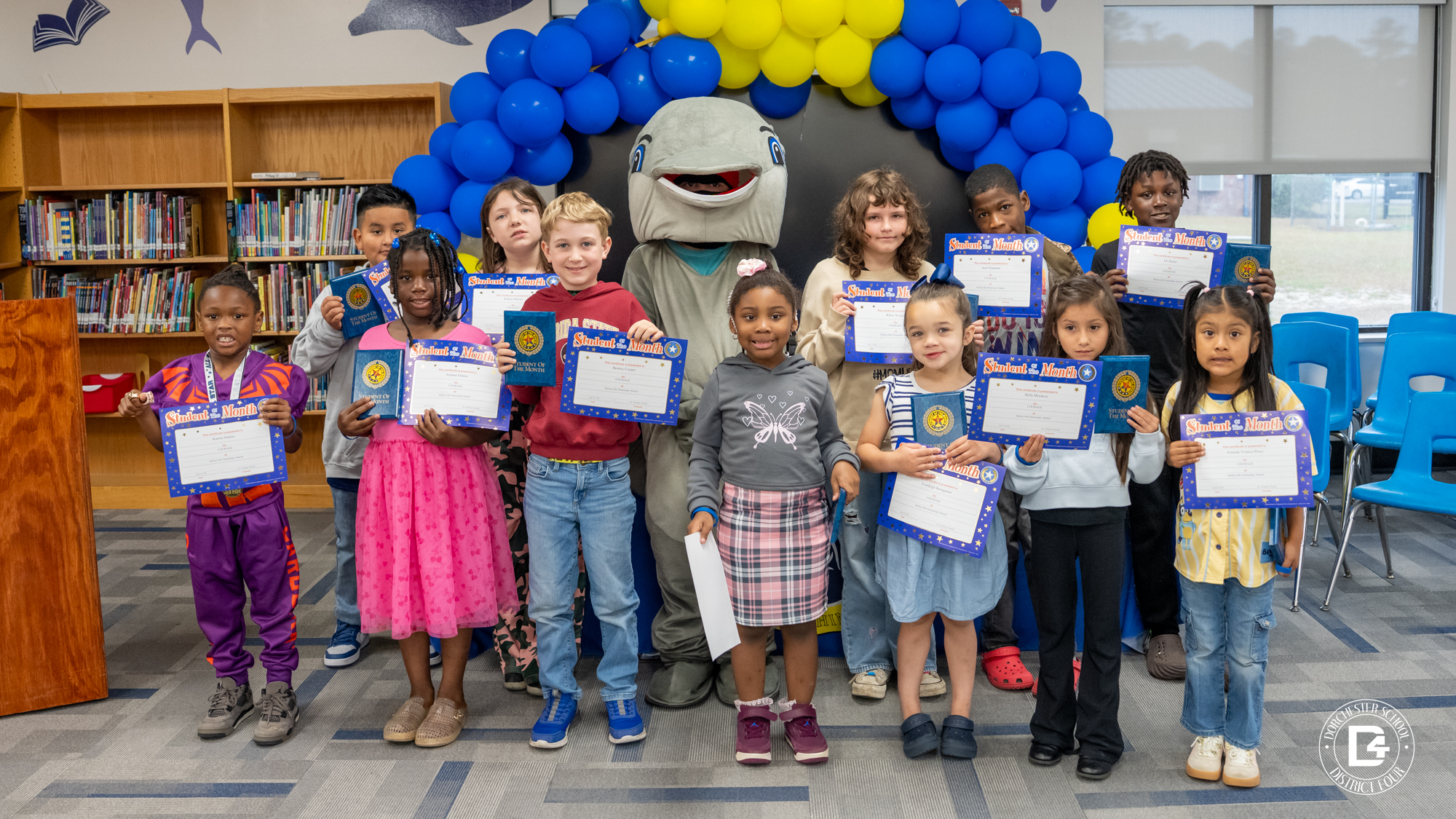 A group of Dorchester School District Four students pose with Student of the Month certificates and folders in front of a blue and yellow balloon display, with the school mascot standing behind them in a library setting.
