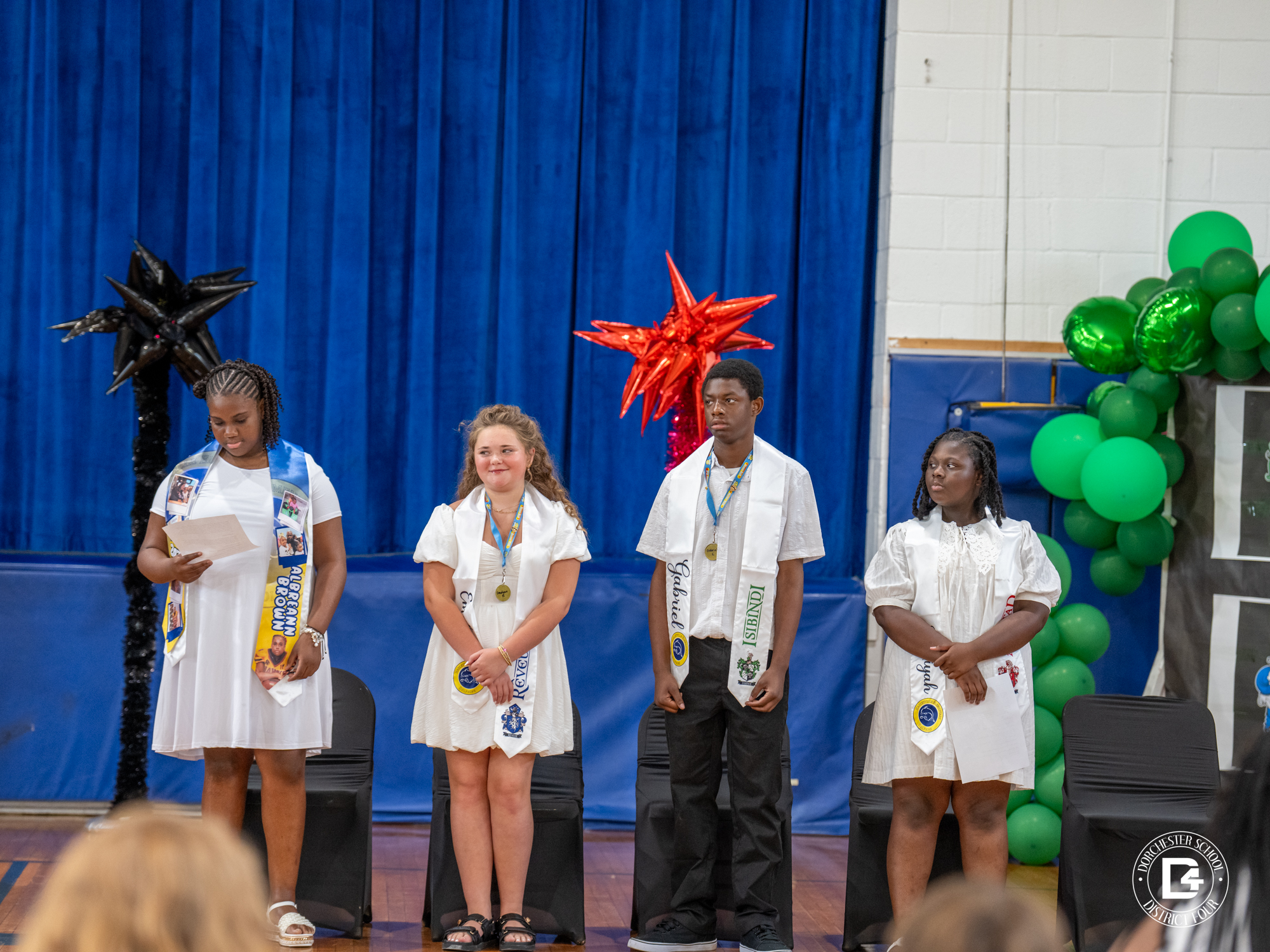 Four students stand at the front of a school gymnasium during a recognition ceremony. Each student wears a white outfit and a decorative stole, with some displaying photos, names, or school-related designs. One student reads from a paper while the others stand attentively. Blue stage curtains, colorful balloon decorations, and chairs arranged behind the students create a festive graduation or awards ceremony setting. Dorchester School District Four branding is visible in the lower-right corner of the image.