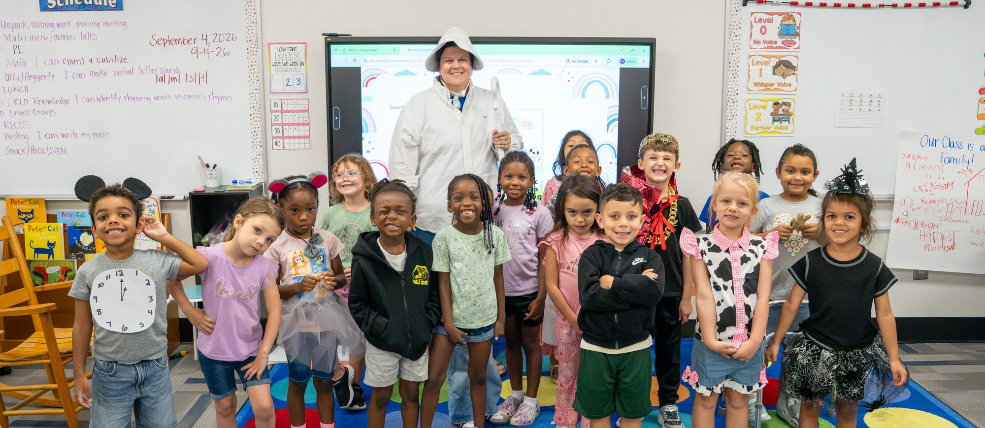A kindergarten class poses for a group photo on a colorful classroom rug with their teacher dressed as Old Mother Hubbard, students wearing various nursery rhyme costumes including a clock, king, cow, and witch.