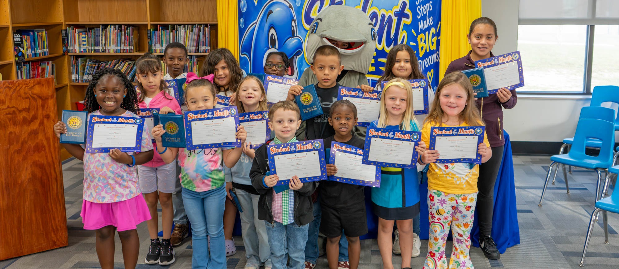 Harleyville Elementary September Character Kids pose in the school library holding their Student of the Month certificates and award folders. The group of smiling students stands in rows in front of a blue and yellow backdrop featuring the school's dolphin mascot and the words "Small Steps Make Big Dreams," with a student wearing the gray dolphin mascot costume standing in the middle of the group.