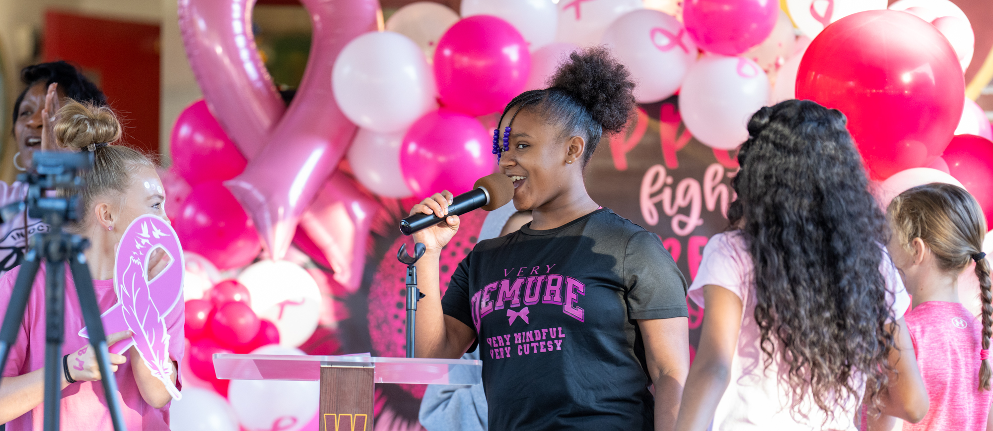 A student at Williams Memorial Elementary School speaks into a microphone during a Pink Out event, surrounded by classmates and a festive backdrop of pink and white balloons and ribbons promoting breast cancer awareness. The podium is labeled “WMES,” and the atmosphere is filled with excitement and encouragement.