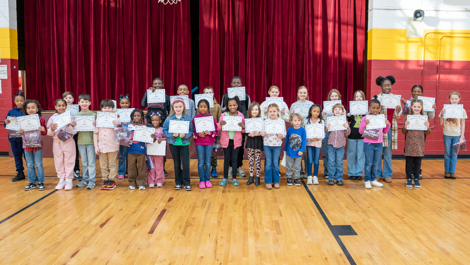 A wide group of Williams Memorial Elementary students stand shoulder to shoulder in a school gym, smiling and holding certificates recognizing them as Character Students of the Month, with a red stage curtain behind them.