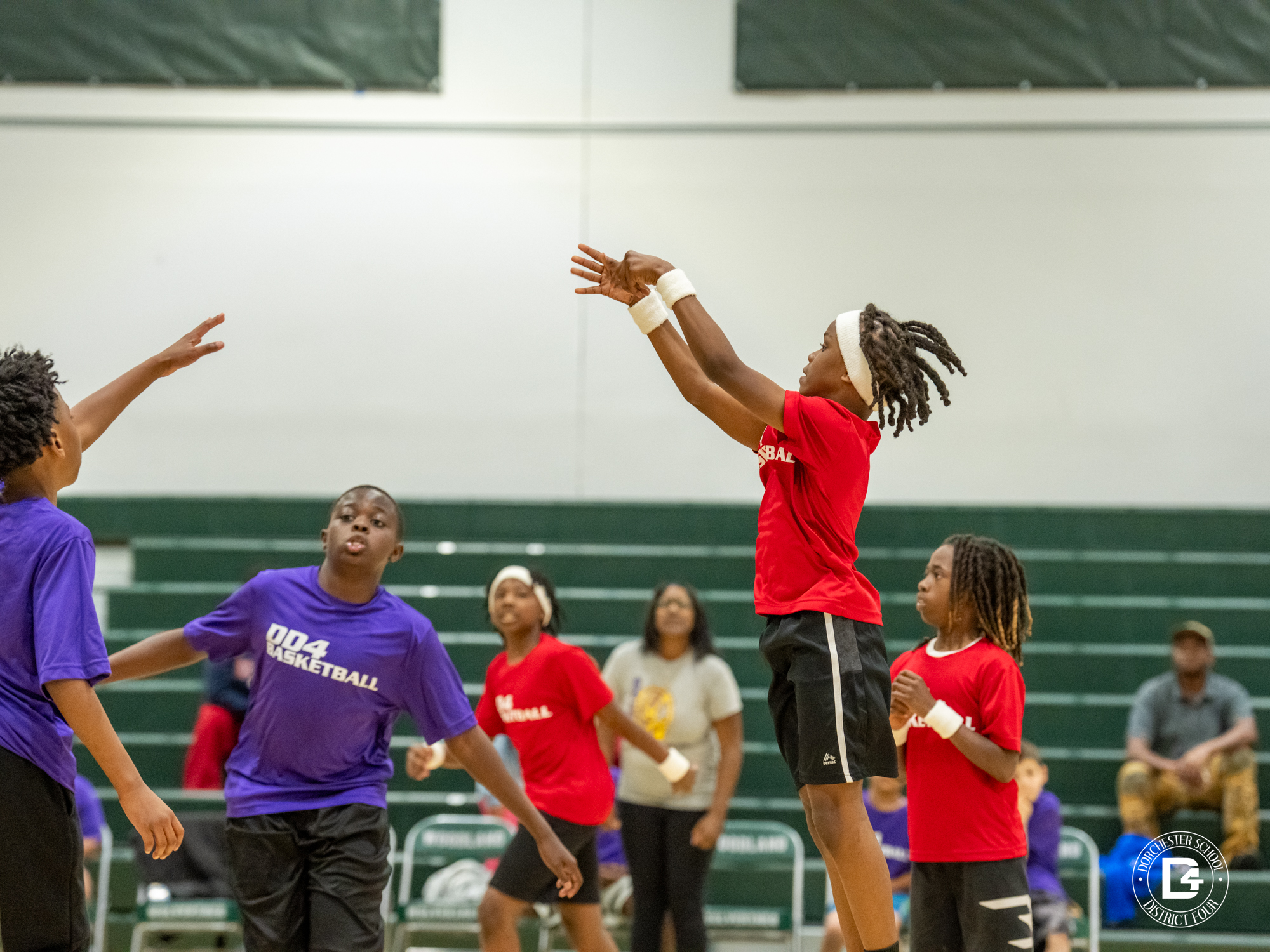 An elementary school student in a red "Basketball" jersey and white headband leaps to release a shot during a Dorchester School District Four youth basketball game. Defenders in purple "DD4 Basketball" jerseys reach up to contest the shot. Additional players in red uniforms and spectators are visible in the background of the green-accented gymnasium. The Dorchester School District Four logo appears in the bottom right corner.