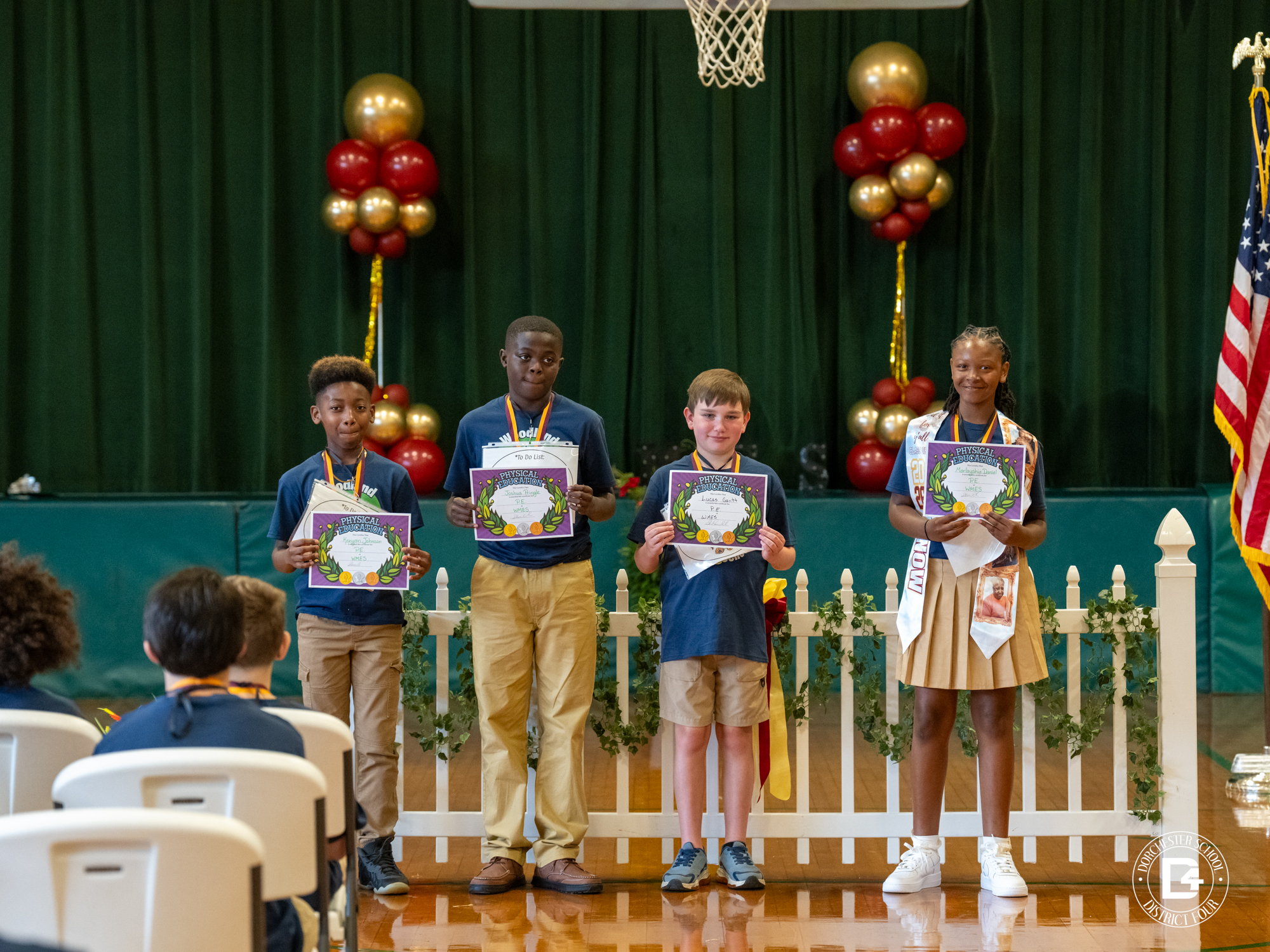 Four students stand on a school gymnasium stage holding Physical Education award certificates during an academic recognition ceremony. The students wear medals and school uniforms, while one student also wears a commemorative stole. A white decorative fence, greenery, red and gold balloon arrangements, and an American flag frame the stage. Seated students watch from the audience as the award recipients pose for recognition. Dorchester School District Four branding appears in the lower-right corner of the image.