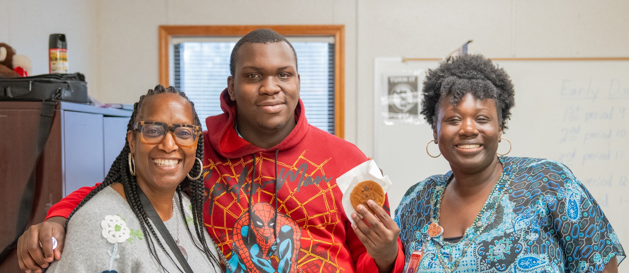 Two staff members and a student holding a juice and a cookie, smiling for the camera at Odyssey. 