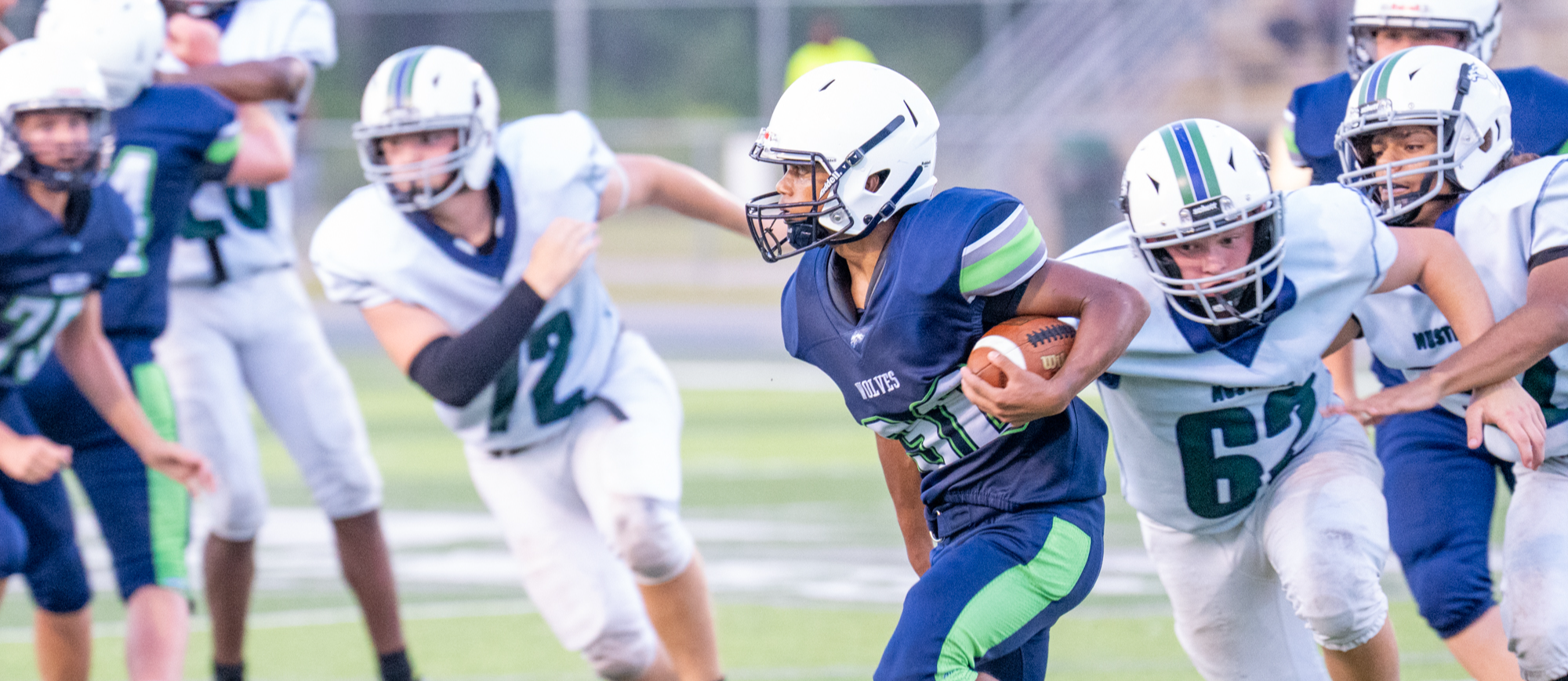 A Woodland Middle School football player in a navy and green Wolves uniform runs the ball down the field during a game. Opposing players in white jerseys and blue pants chase closely behind as the player looks for an opening. The action takes place on a well-lit field, capturing the intensity and focus of the play.