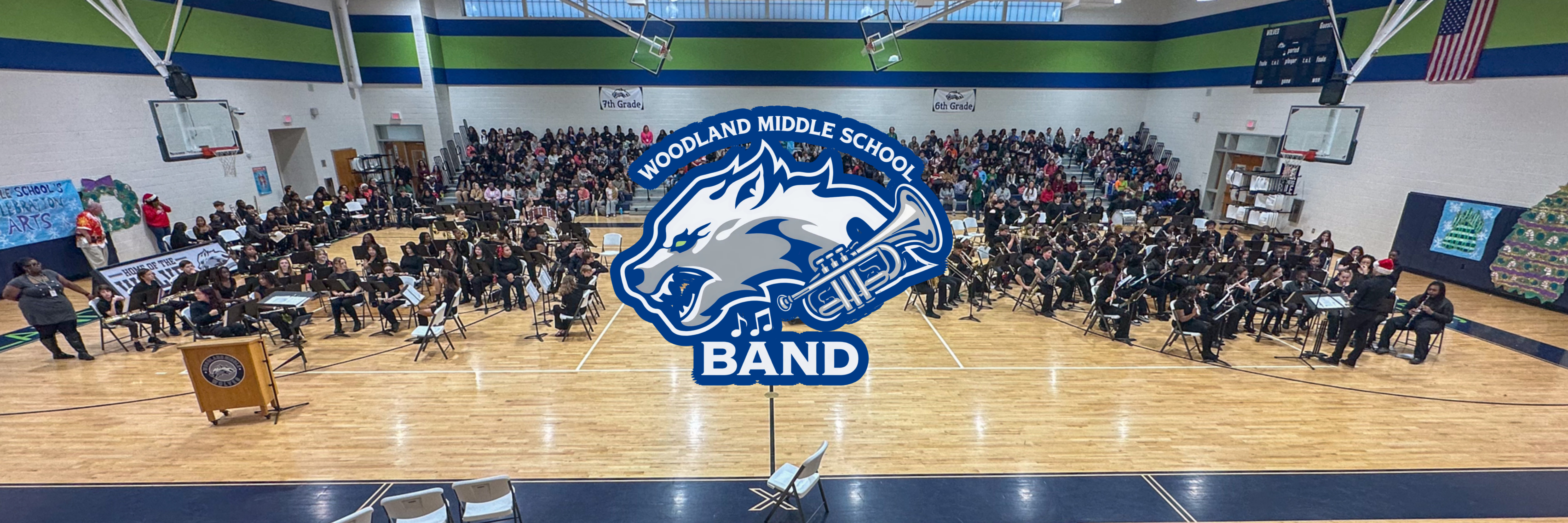 Wide panoramic photo inside a school gym showing the Woodland Middle School band seated on the court with instruments ready to perform, a large audience filling the bleachers behind them, holiday decorations on the walls, and a Woodland Middle School Band wolf logo overlaid at center court.