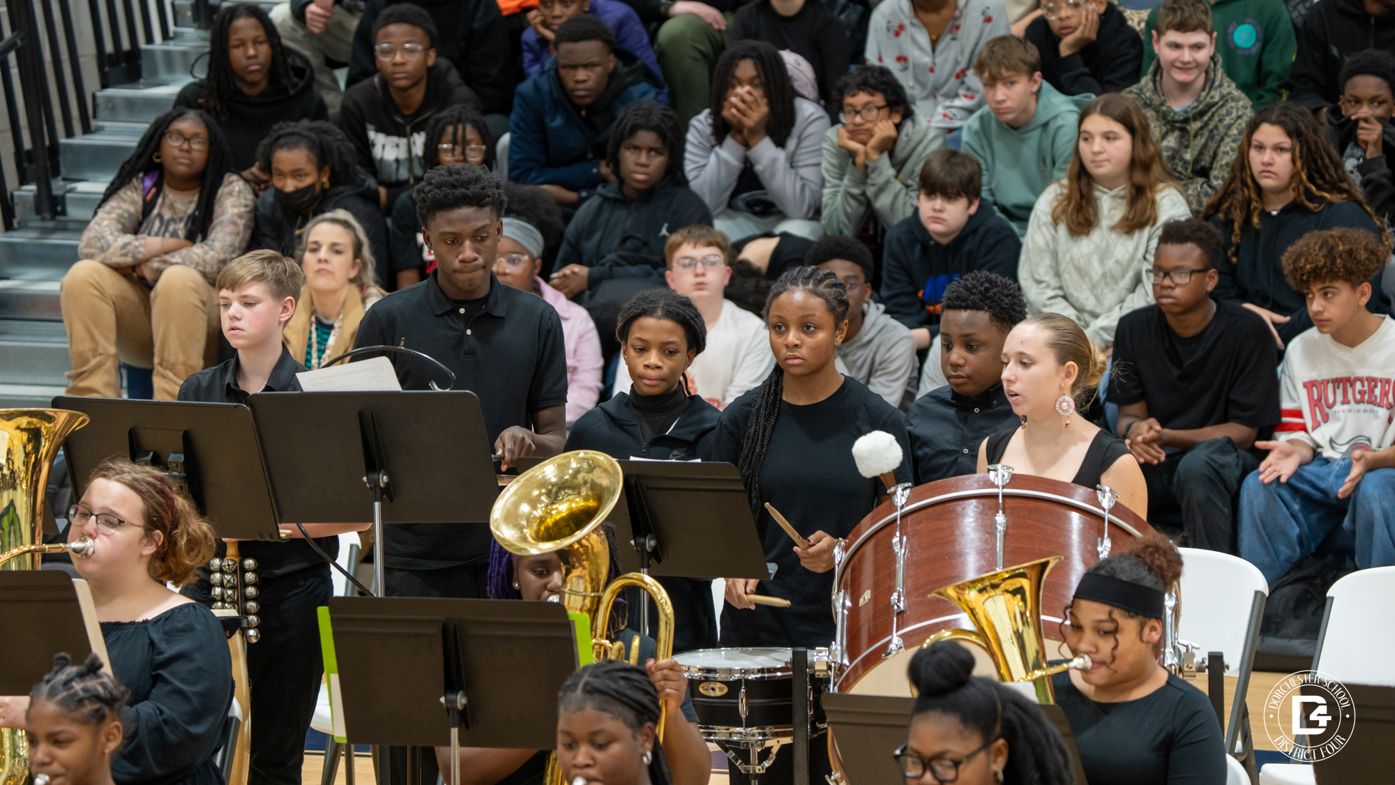 Students inThe percussion section of the Woodland Middle School band performs behind the ensemble, with a large bass drum centered and students focused on the conductor.the Woodland Middle School band perform on saxophones and brass instruments, seated in rows with music stands during a gymnasium concert.