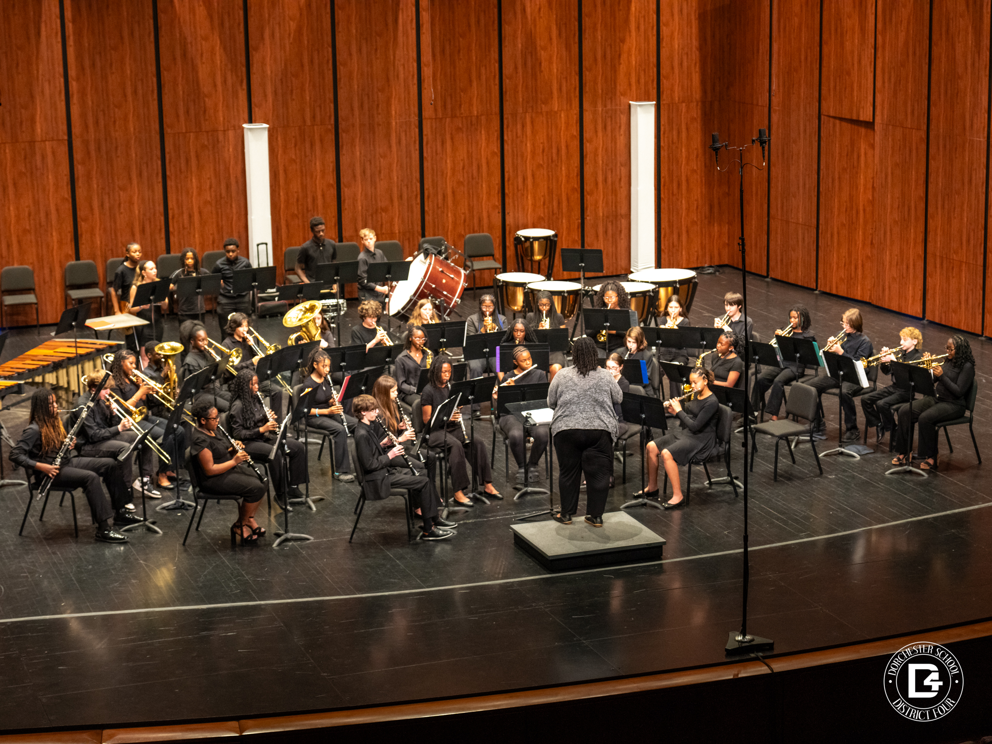 Woodland Middle School concert band performs on stage in a concert hall, dressed in black attire, seated in sections with instruments including flutes, clarinets, trumpets, trombones, and percussion. The band is led by director Ms. Yolanda Bennett, who stands on a podium conducting in front of the group, with wooden acoustic panels behind them and stage microphones set up.