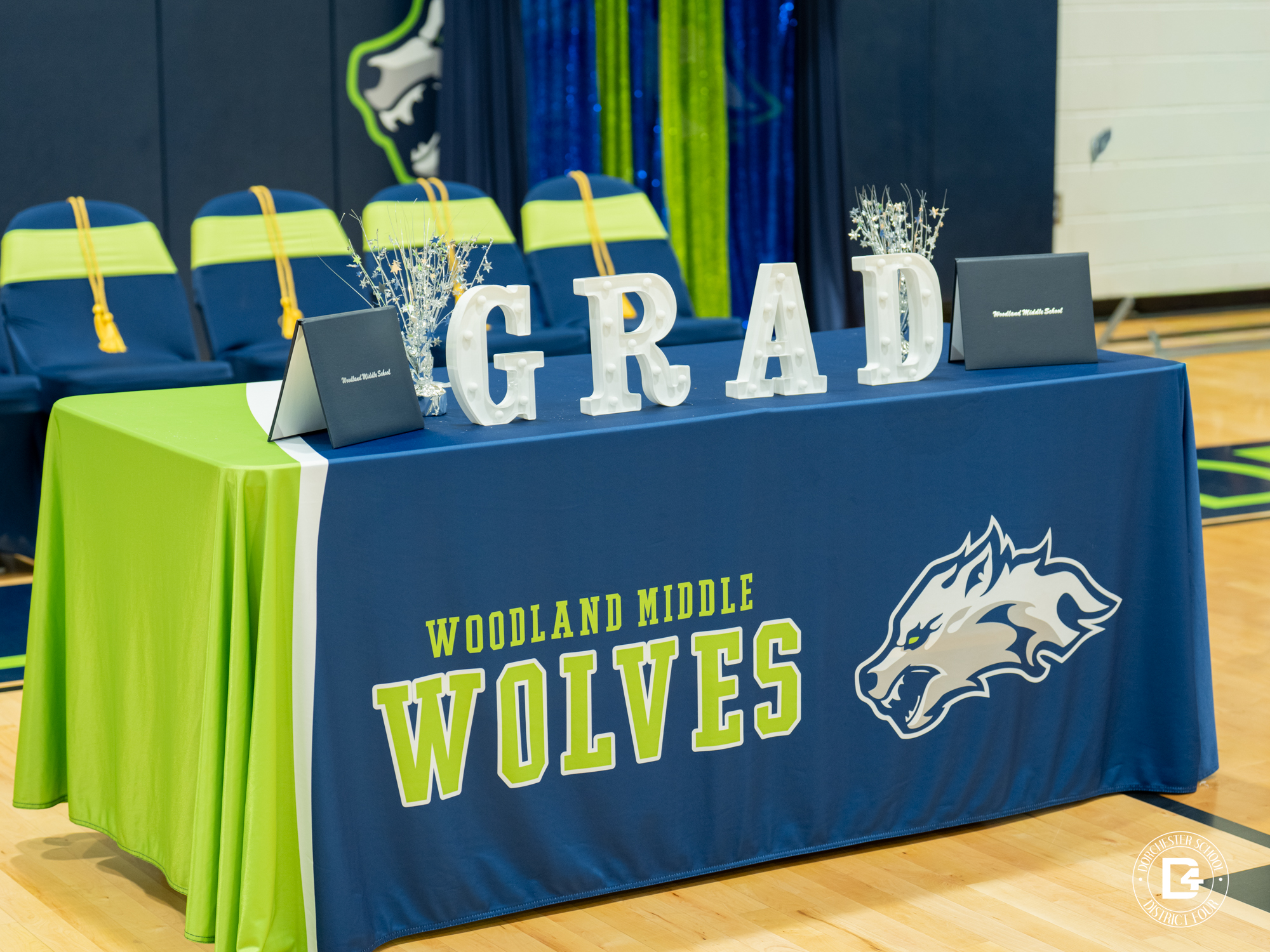 A decorated table at Woodland Middle School displays illuminated letters spelling “GRAD,” diploma covers, and silver centerpieces. The table is covered in blue and lime green linens featuring the Woodland Middle Wolves logo.