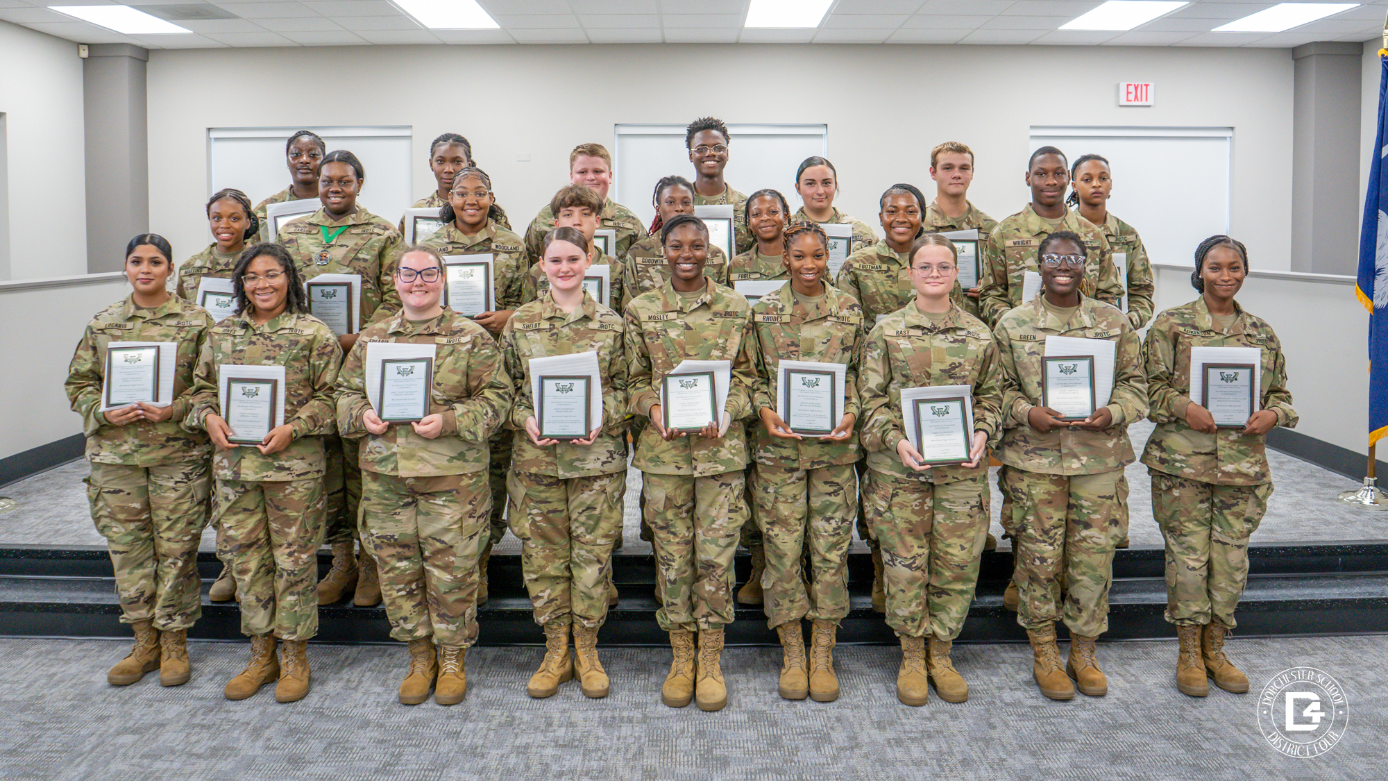 A large group of Woodland High School JROTC cadets in camouflage uniforms pose on steps inside the board room, each proudly holding a wooden plaque of recognition.