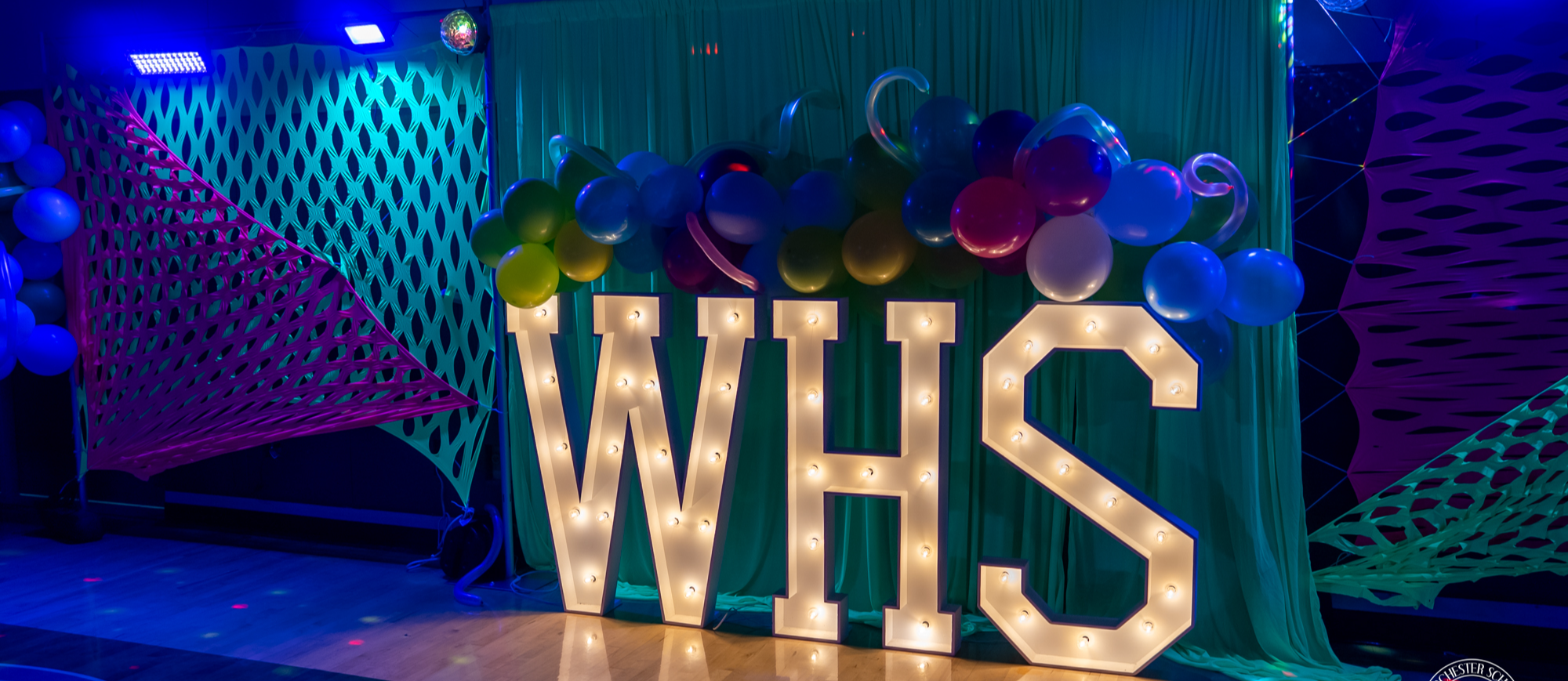 A decorated event backdrop features large illuminated letters spelling "WHS" on a gym floor. Colorful balloons are arranged across the top, with green fabric draping behind and neon pink and green netting stretched on either side. Blue and purple stage lighting creates a vibrant glow, giving the setup a festive and celebratory atmosphere.