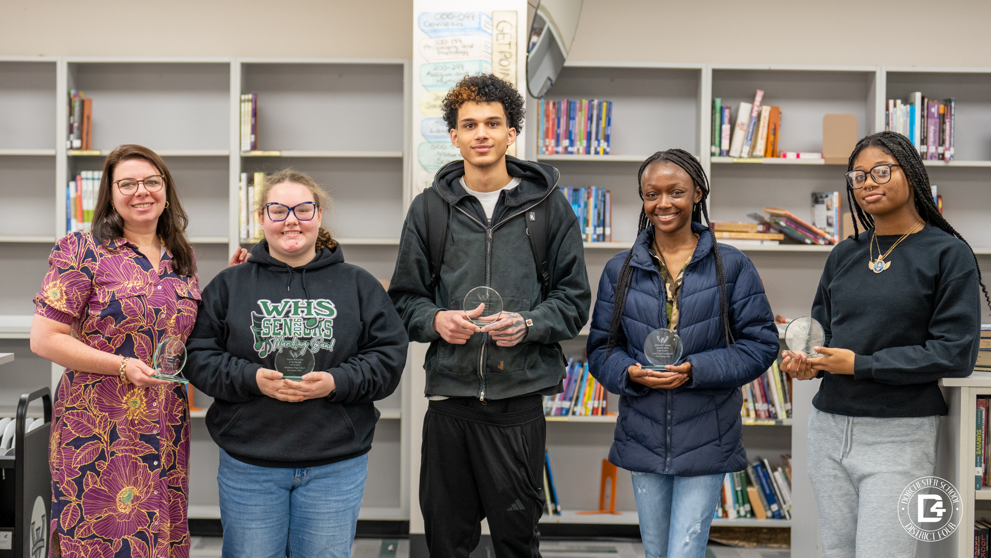 Woodland High School Wolverines of the Month stand in the media center holding recognition awards, posing in front of bookshelves with a staff member as part of the monthly student recognition program.