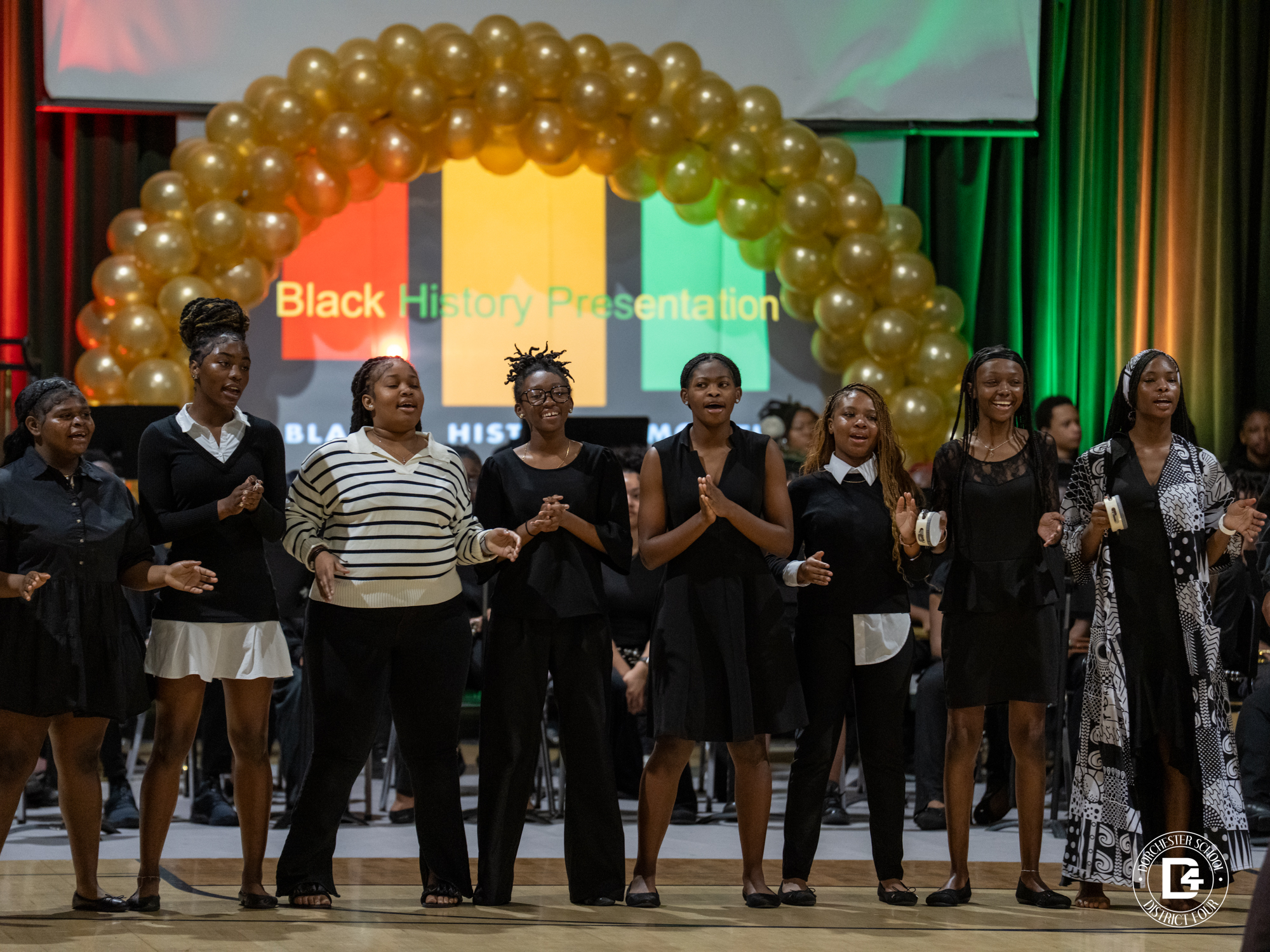 Eight female students dressed primarily in black stand across a stage singing during a Black History Presentation. A large gold balloon arch frames them, and a screen behind displays the words “Black History Presentation” with red, yellow, and green colors. Additional students with musical instruments are seated in the background, and a Dorchester School District Four logo watermark appears in the bottom right corner.