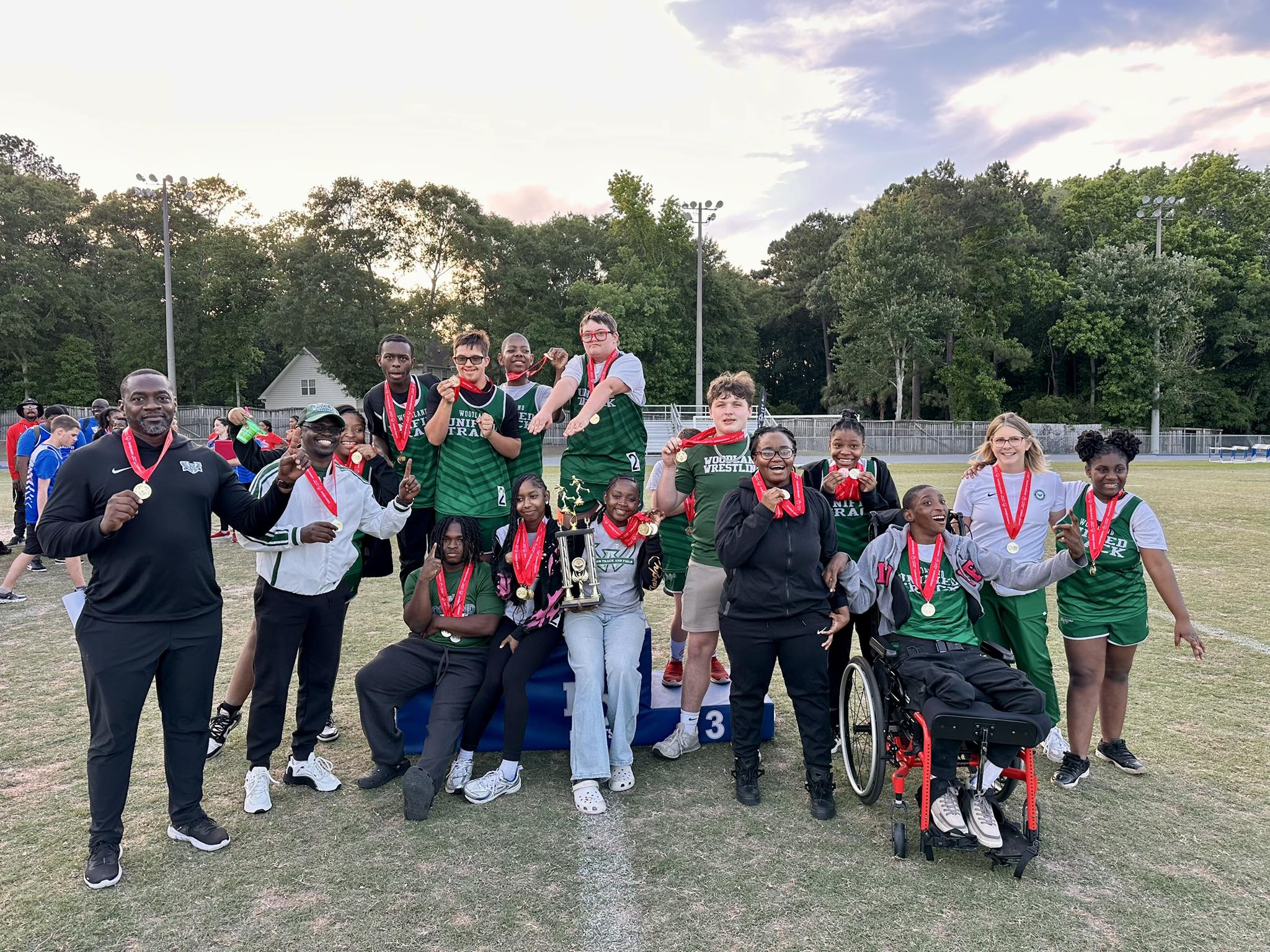 Woodland High School Unified Track &amp; Field team members and coaches pose on the track after winning Lower State, smiling and holding medals, a trophy, and a plaque.