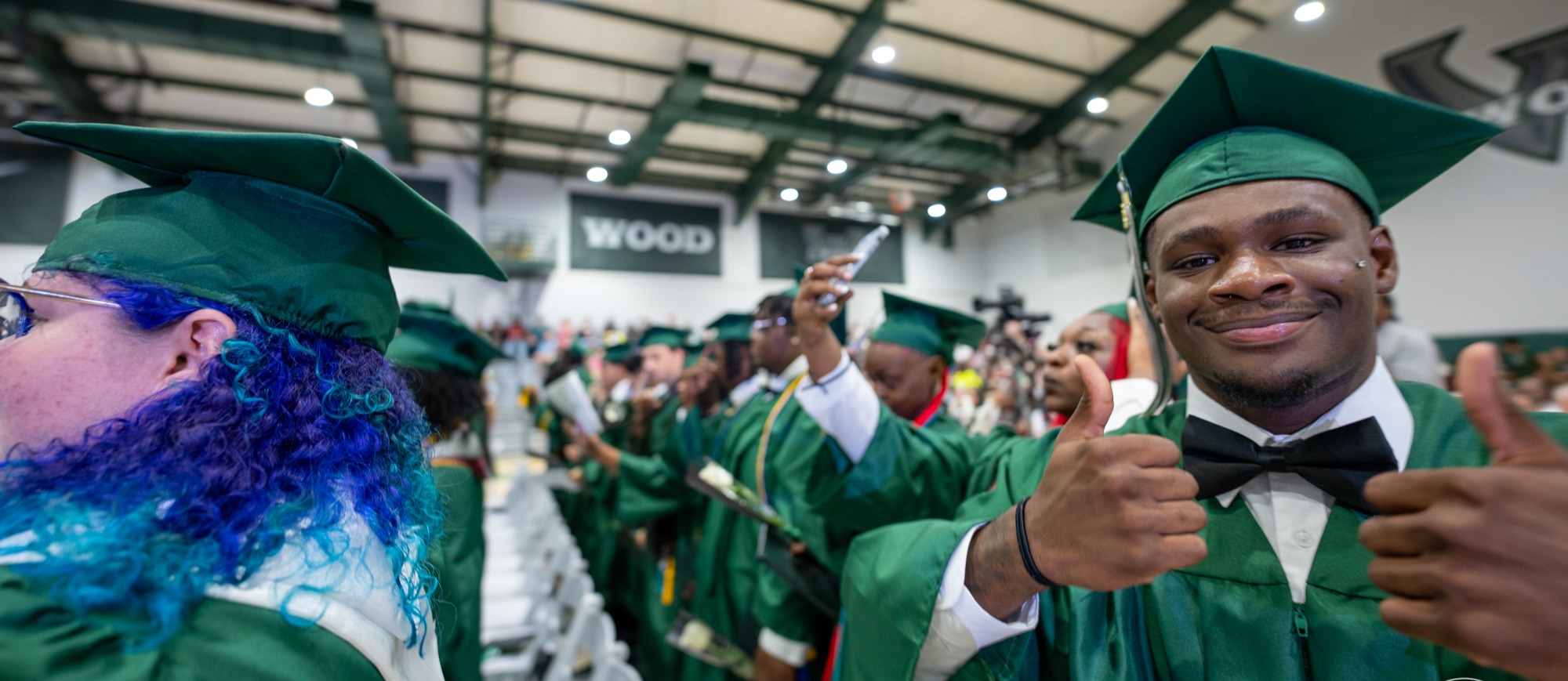 Woodland High School graduates stand together during commencement exercises inside the school gymnasium. Students wearing green caps and gowns smile, wave, give thumbs-up, take photos, and celebrate with classmates as family members and guests watch from the stands. Many graduates wear honor cords, medals, and stoles, reflecting academic achievements and involvement in school organizations. The atmosphere is energetic and joyful as the Class of 2026 prepares to receive diplomas and mark the culmination of their high school journey.
