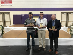Three men proudly display their awards in a gymnasium, celebrating their achievements in a vibrant atmosphere.