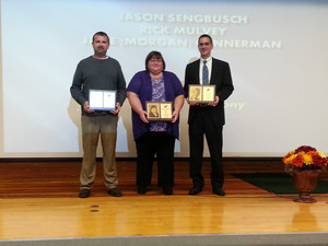 Three individuals on stage proudly holding their awards, celebrating their achievements in front of an audience.