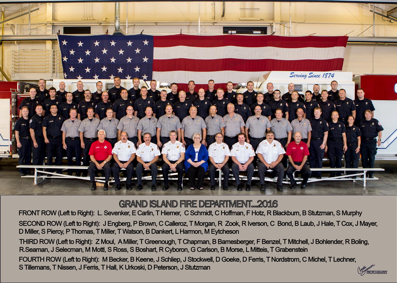 A group of firefighters posing together, proudly displaying their 2016 team photo in front of a flag, with names listed at the bottom.