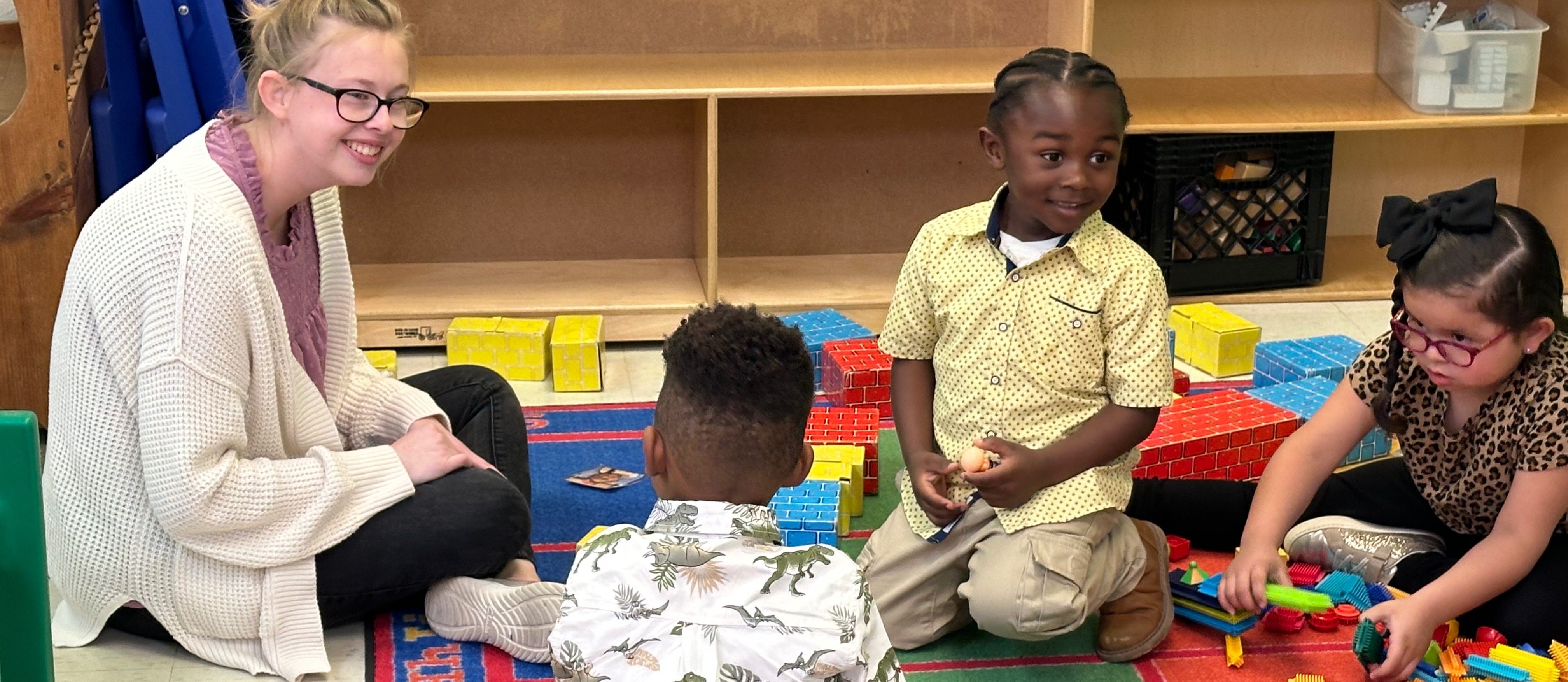 A teacher is seated on the floor with her students