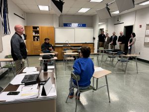 A classroom scene with students at desks and a man teaching at the front