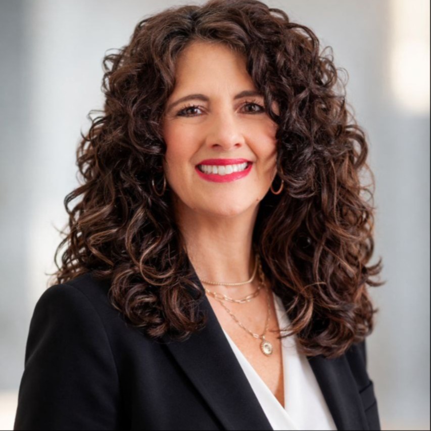 Head shot of Gretchen Montgomery, Elemeentary Math District Instructional coach. She is wearig a black blazer and a white v neck shirt. She has long brown curly hair and brown eyes.
