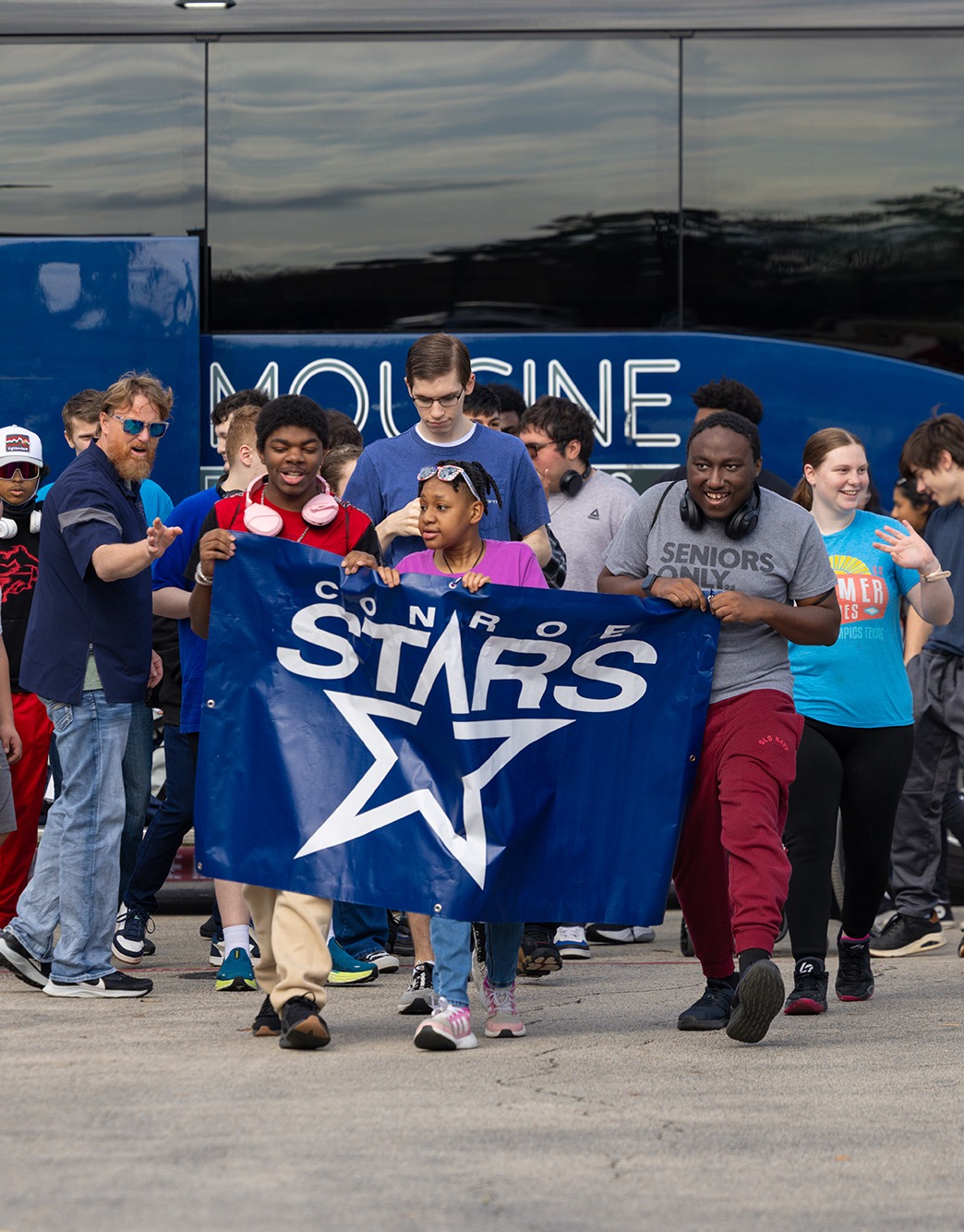 Large group of students in front of a charter bus. Three of them are carrying the Conroe Stars banner.