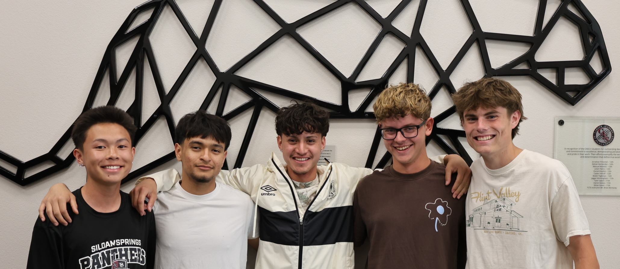 Five young men pose in front of a wall with a geometric black cat sculpture.