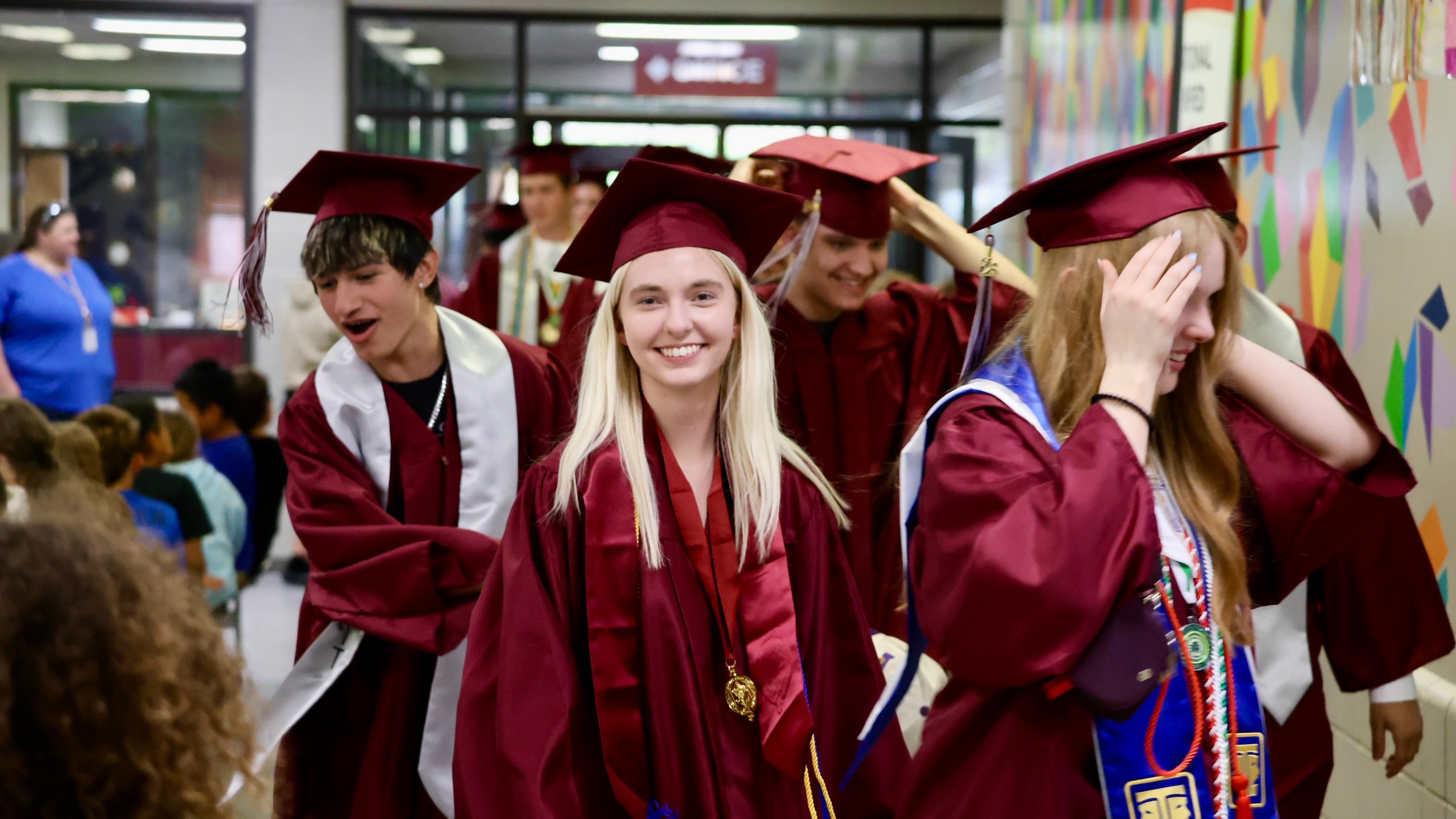 A group of students in maroon gowns and caps are smiling and walking through a hallway.