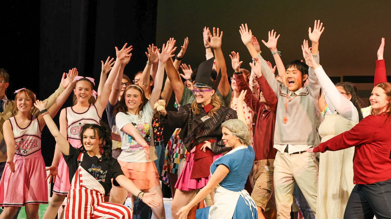 Group of performers in various costumes and outfits on stage, hands raised, with a speaker on the floor.