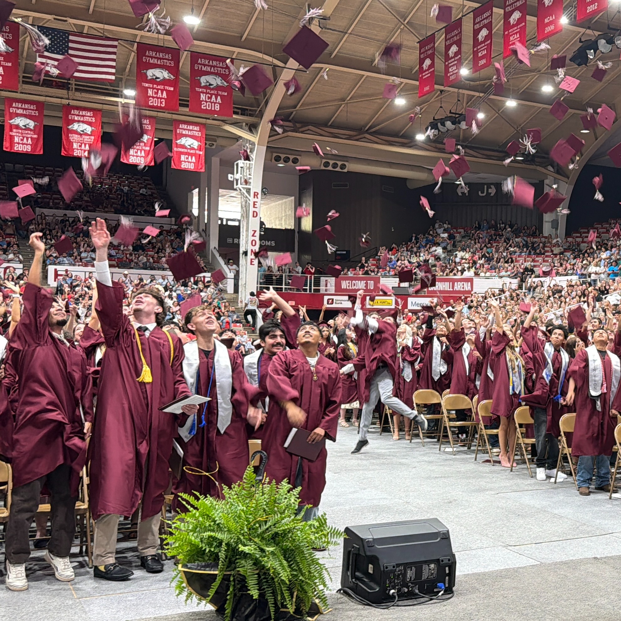 Graduates in maroon robes throw caps into the air at an outdoor ceremony. A woman stands at a podium.