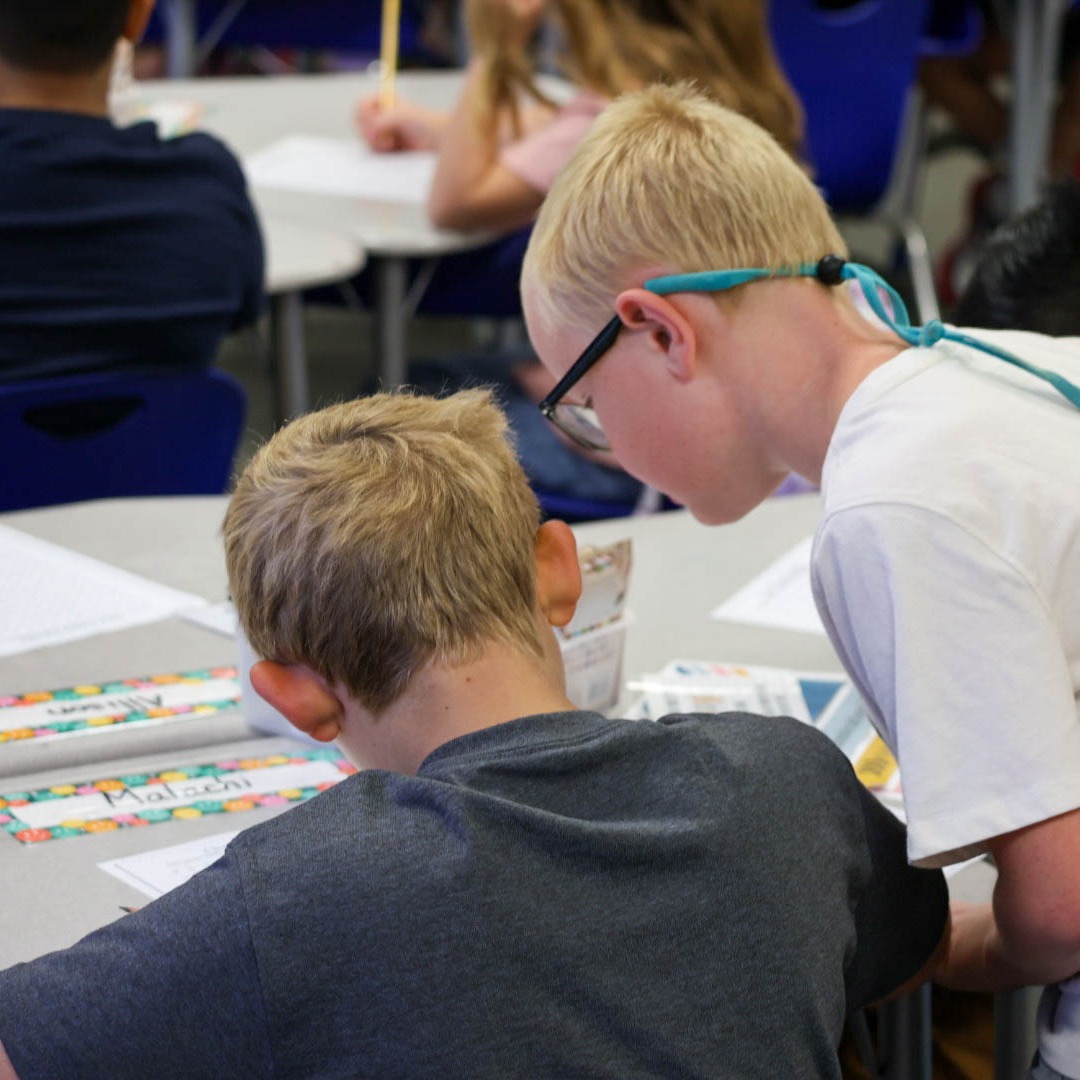 Students in a classroom; one boy in glasses leans over a table, looking at another boy's paper.