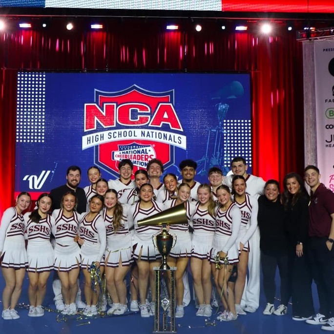 Cheerleaders stand with a trophy on a stage, wearing uniforms. Behind them, a banner reads "NCA NATIONAL CHAMPIONS."