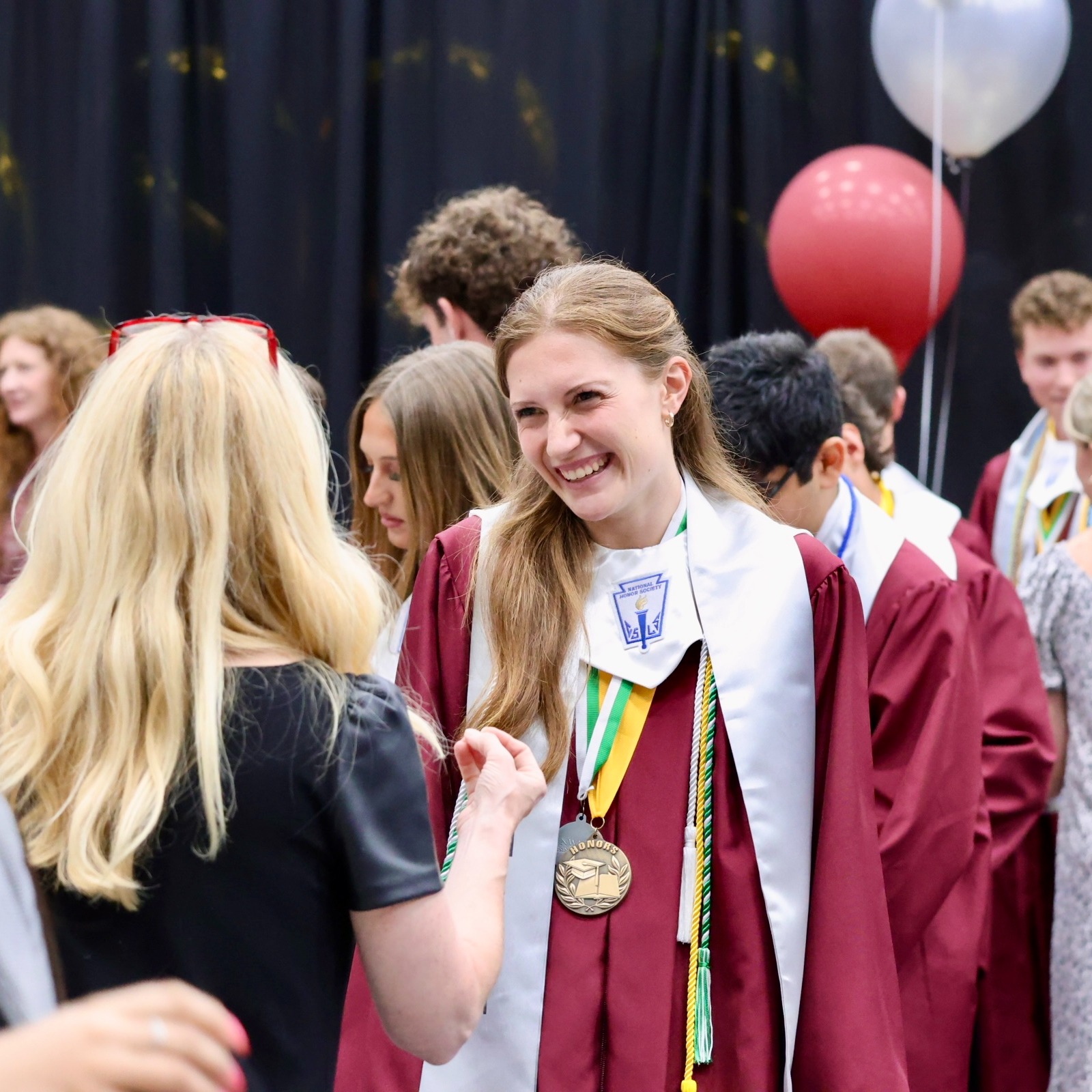 Graduates in robes, some with medals, standing close together. Background shows a black curtain and balloons.