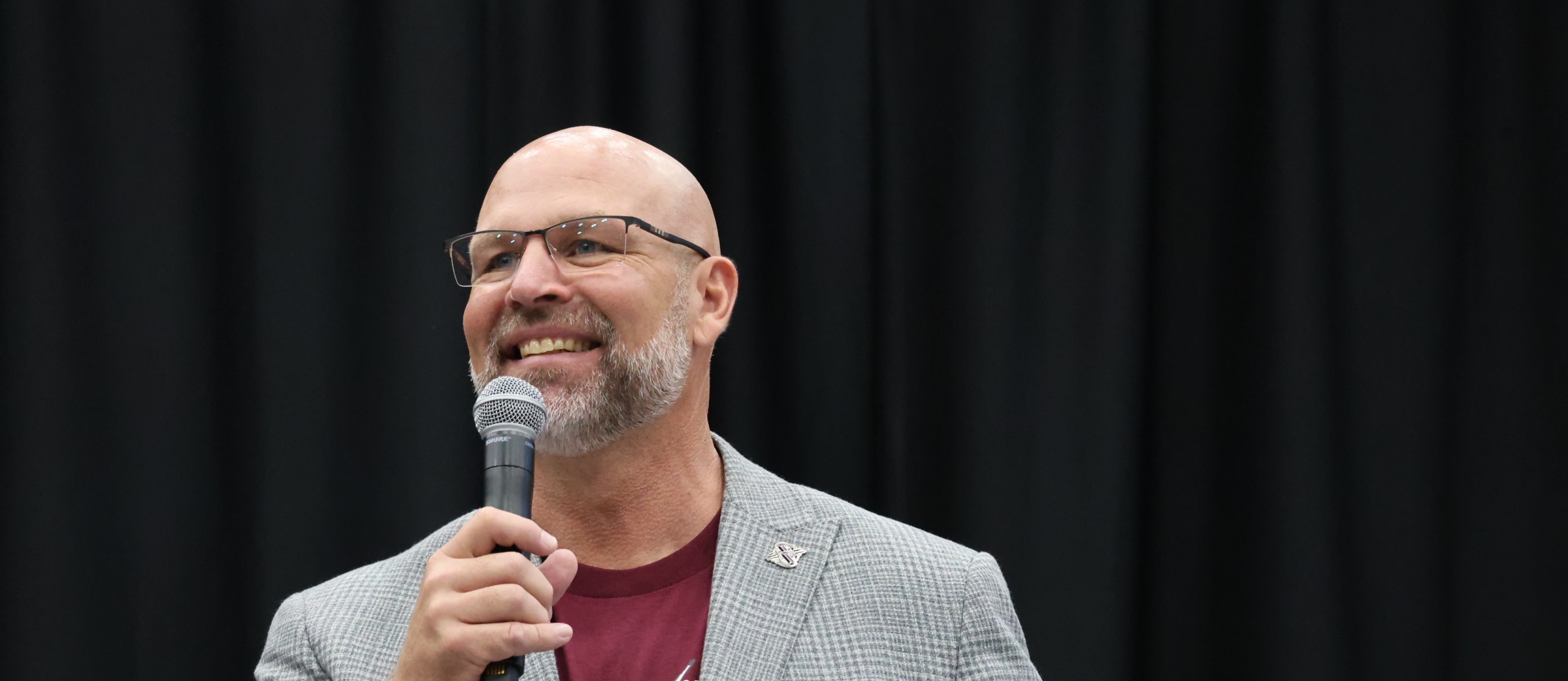 Man with glasses, gray jacket, and red shirt, standing and smiling, holds a microphone. Black curtain behind.