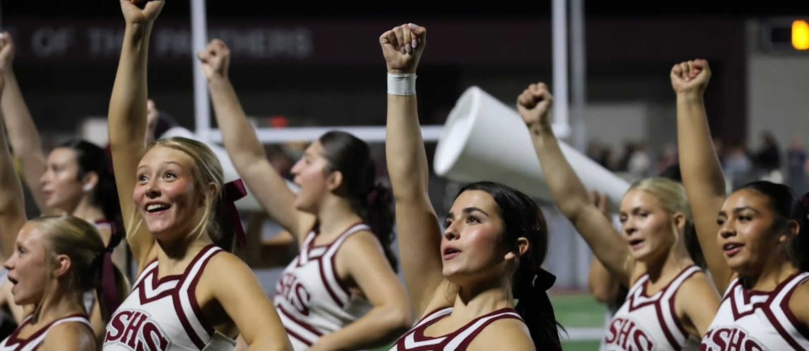 Cheerleaders in white and maroon uniforms with raised arms, two holding megaphones, and a dark background.